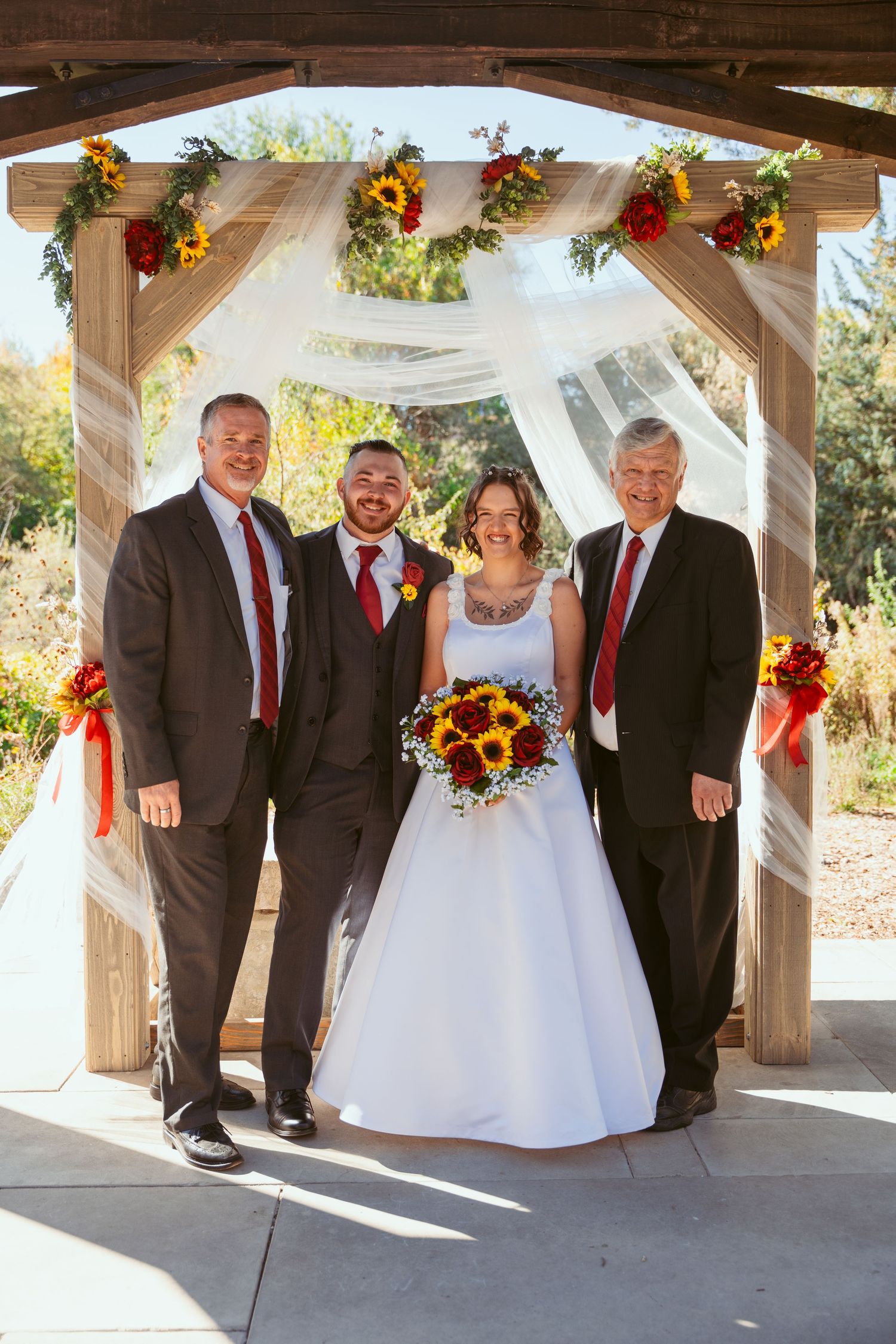 Wedding party poses under wooden pavilion decorated with white fabric and sunflowers in outdoor setting.