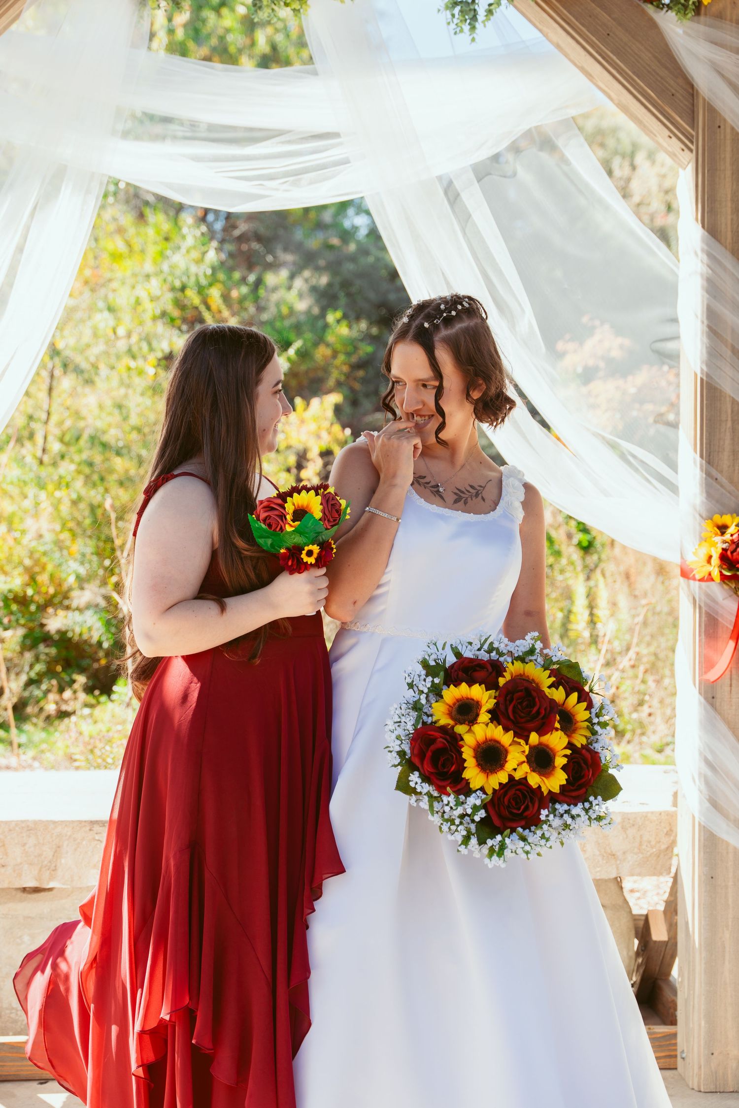 Wedding party members share joyful moments with sunflower bouquets under white draped pergola.