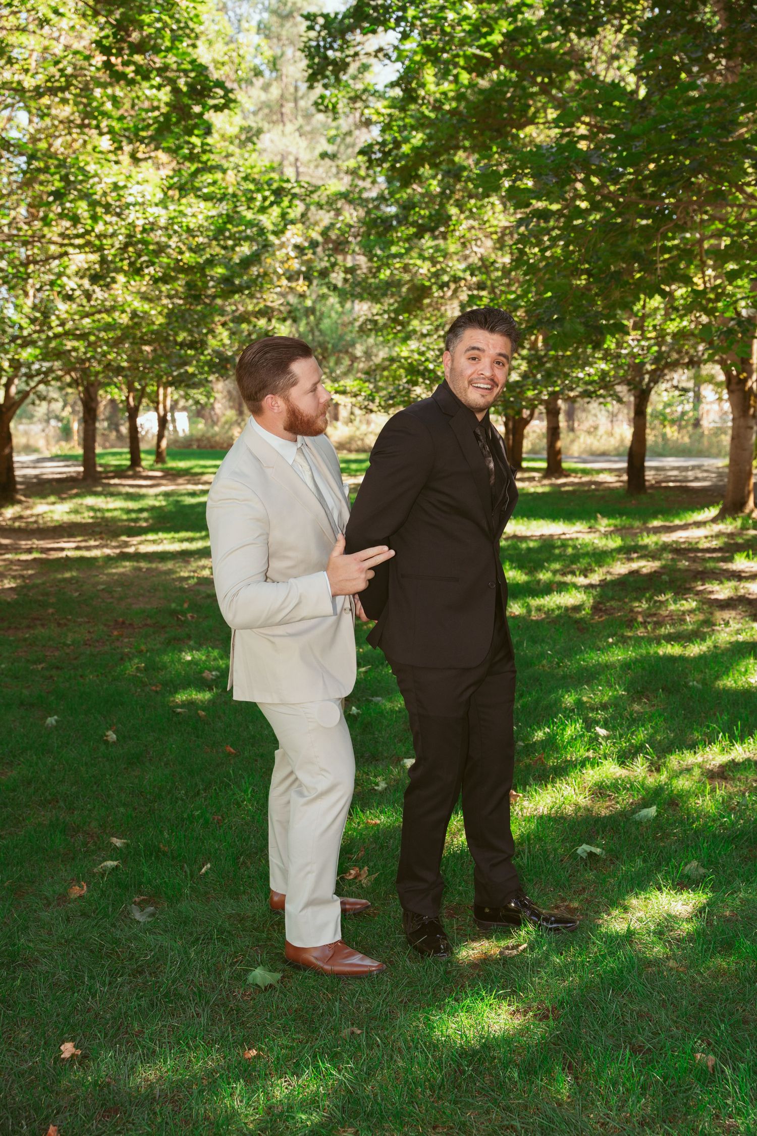 Two people in contrasting formal wear interact playfully on grass under a canopy of trees.