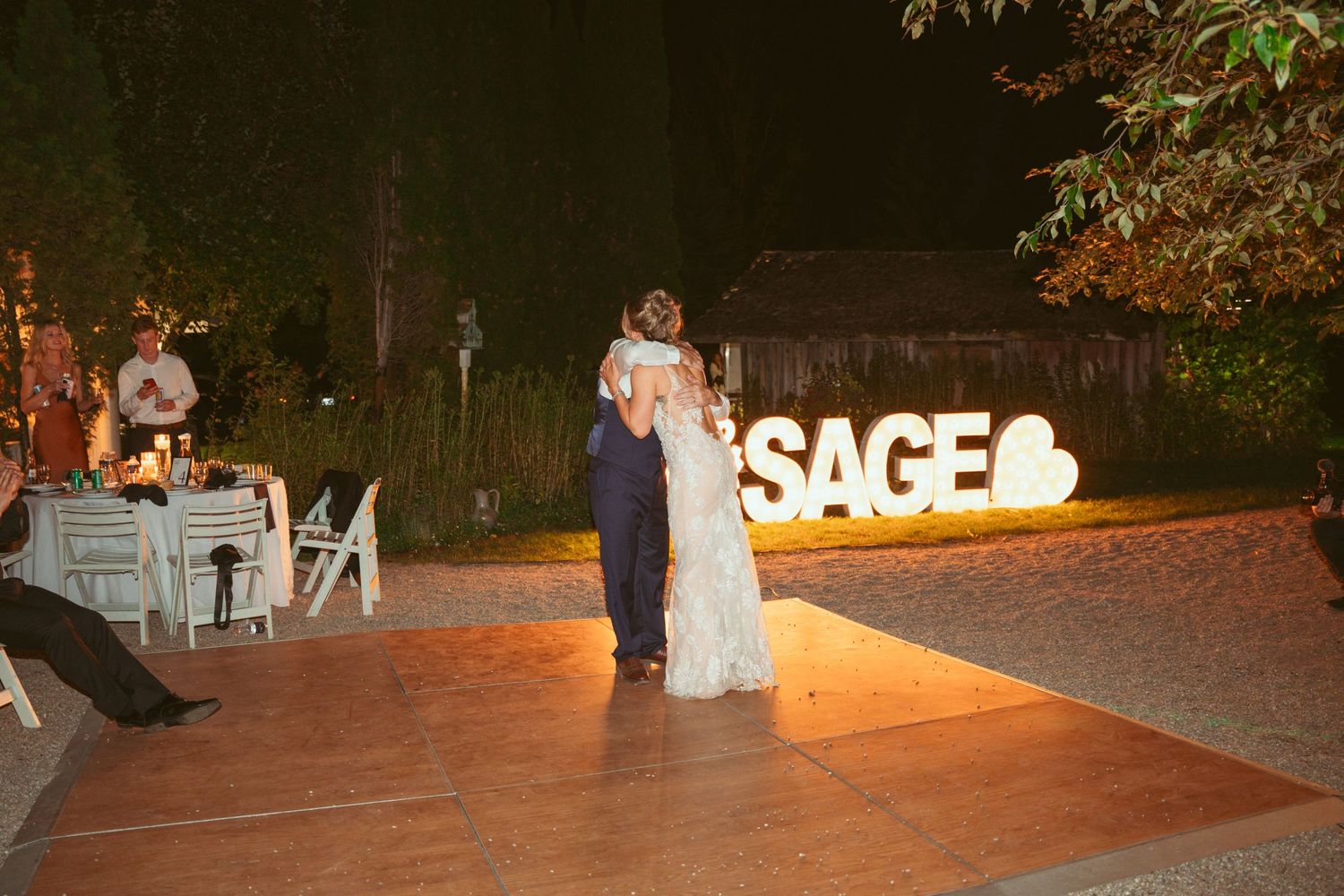 A couple shares their first dance on an outdoor dance floor with illuminated SAGE signage in the background.
