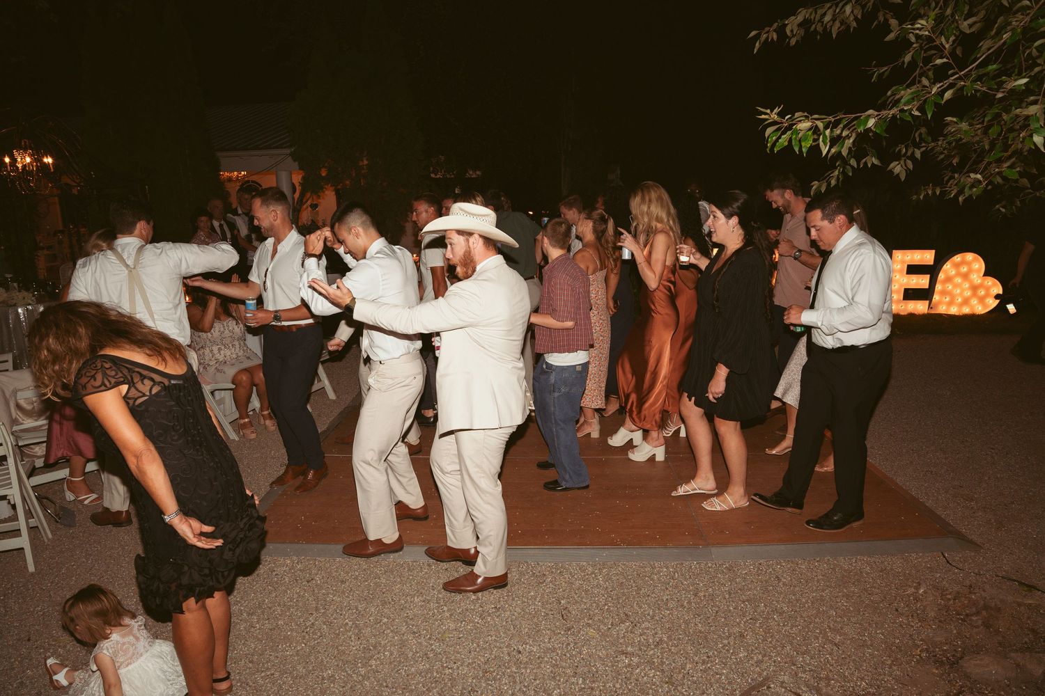 Wedding guests dancing in a line at an outdoor evening reception.