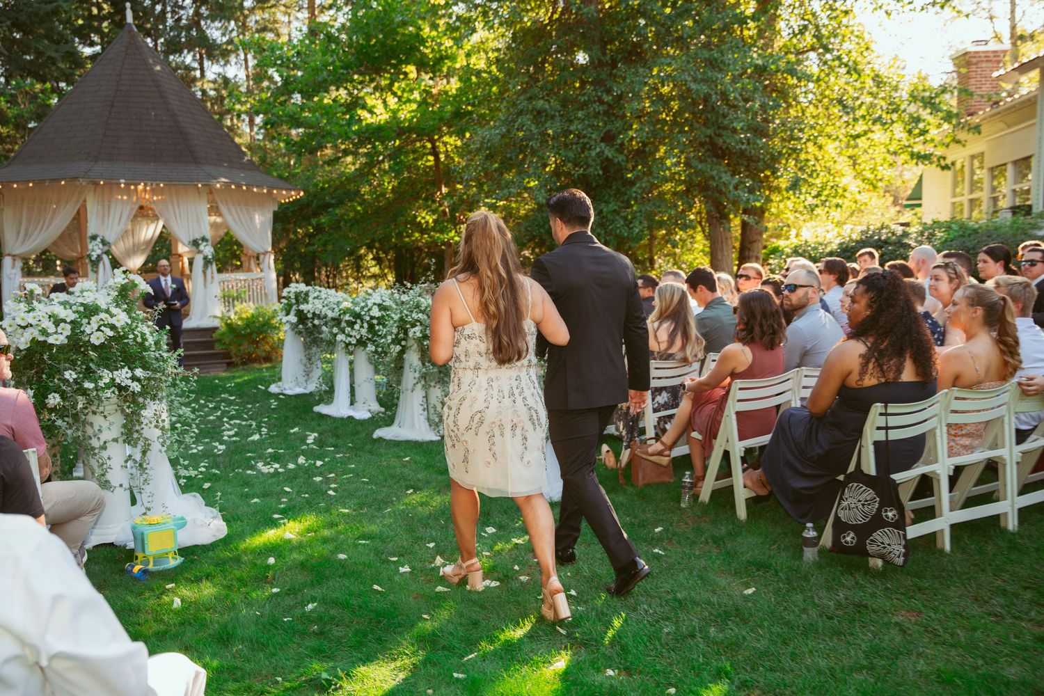 Couple walks down aisle after ceremony in garden setting with white chairs and decorated gazebo in background.