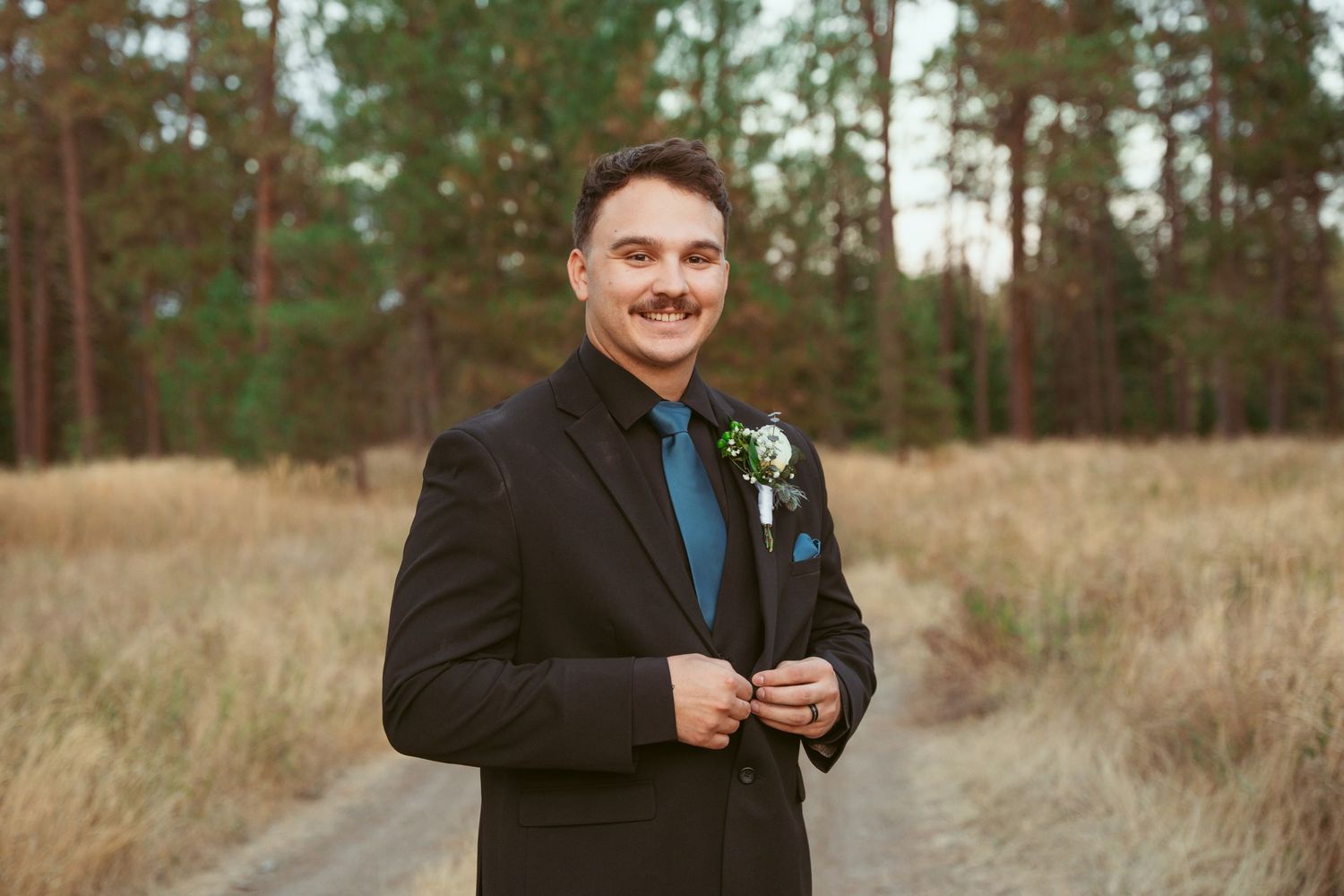 A person in a black suit and blue tie smiles while standing in a rustic outdoor setting.