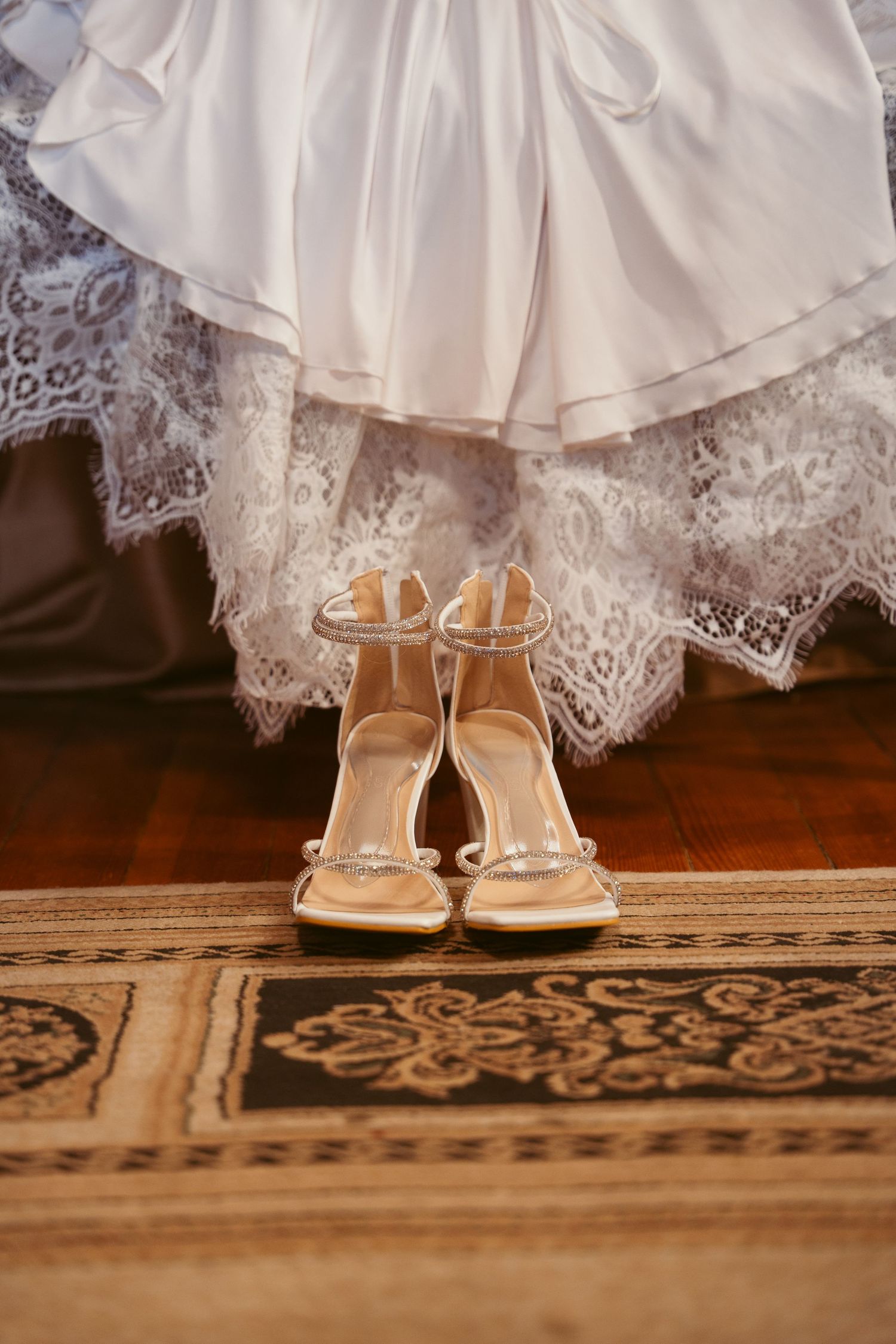 Delicate lace wedding dress hem and nude strappy heels on a patterned rug.