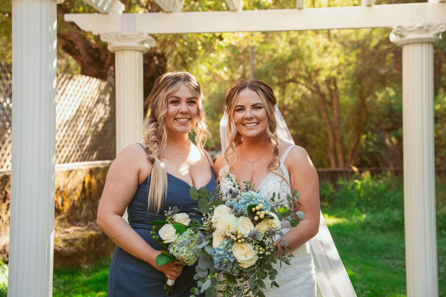 Two wedding attendants pose with matching blue and white floral bouquets under white pergola.