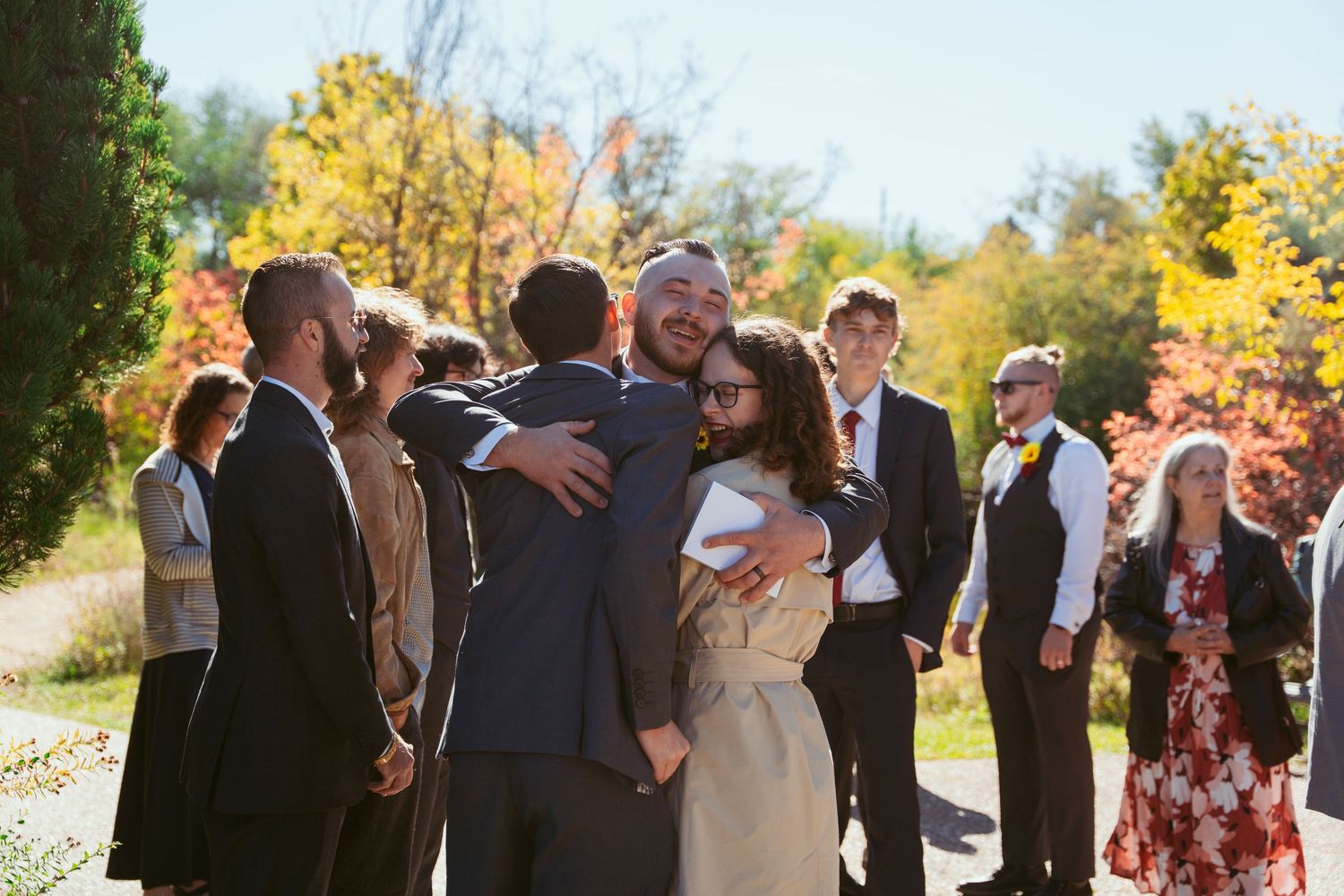 Wedding guests gather outdoors on a sunny autumn day surrounded by fall foliage.