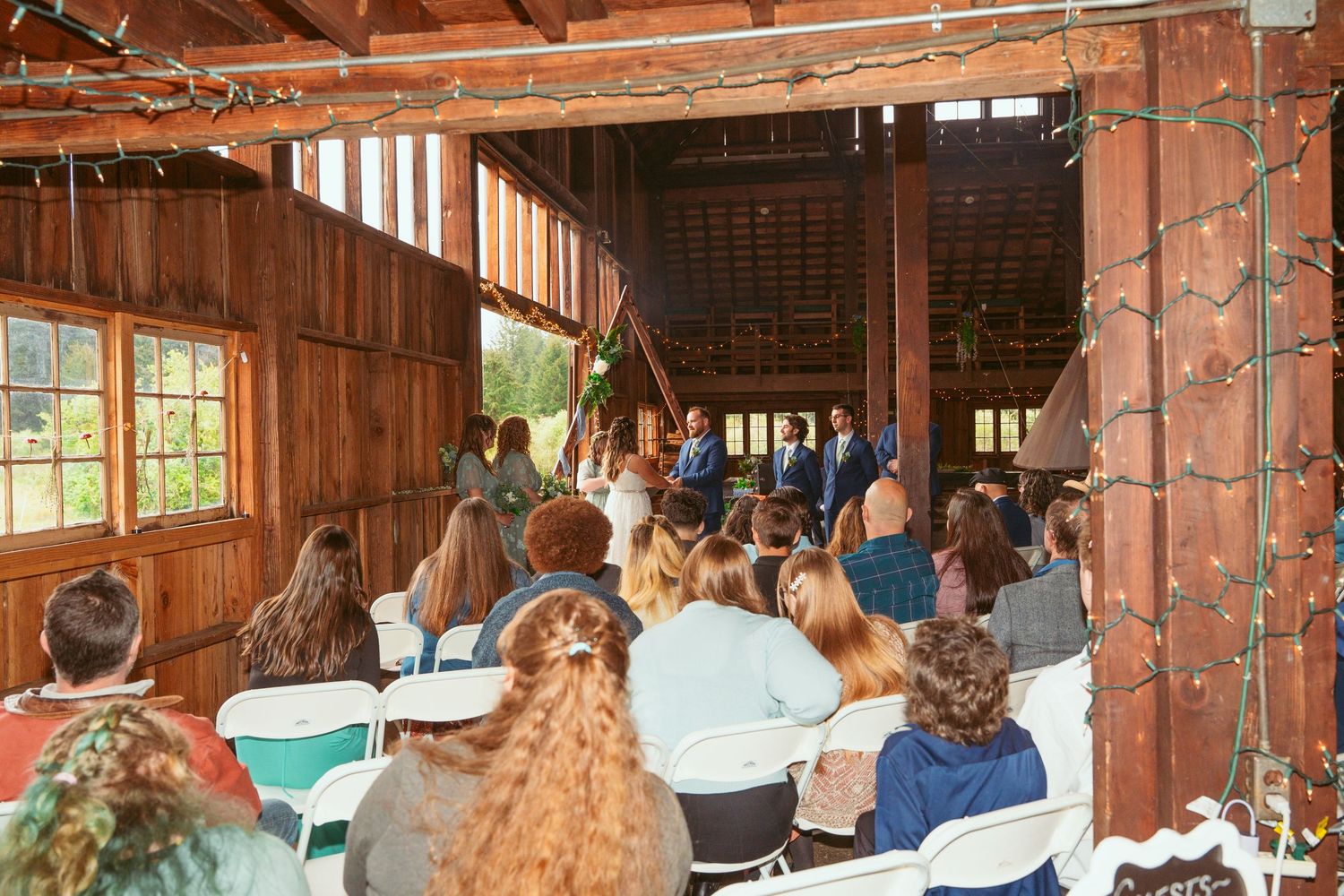 Wedding guests watching ceremony from wooden chairs in rustic barn setting.