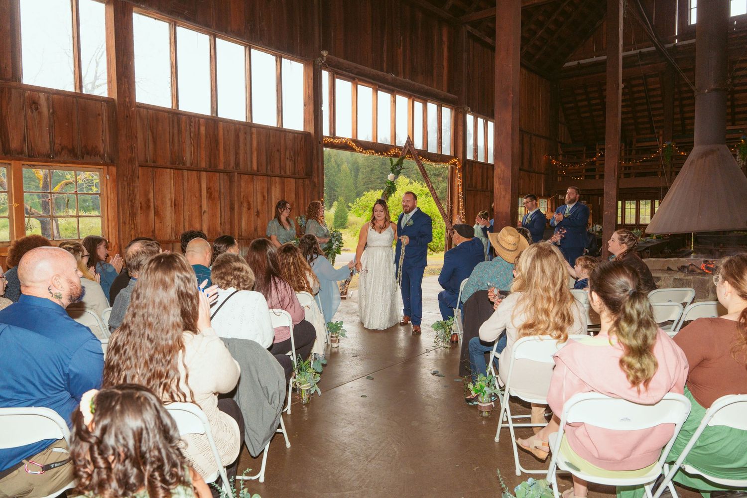 Wedding guests seated in rows of white folding chairs inside a rustic wooden barn venue.