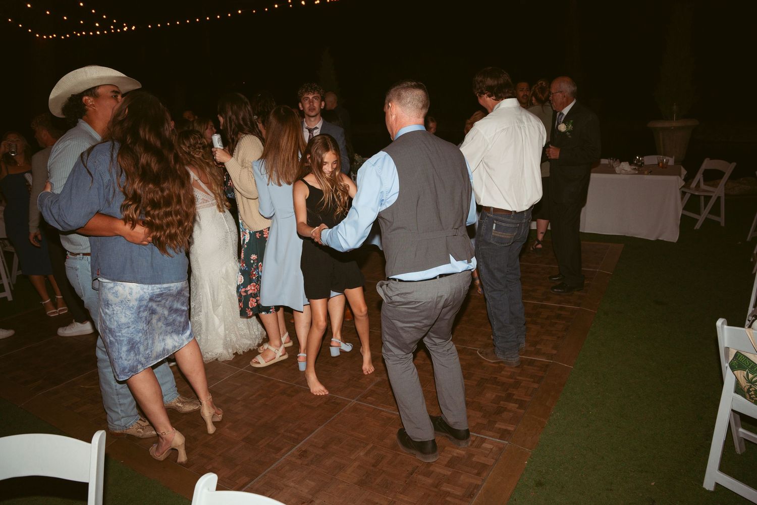 Wedding party dancing on an outdoor patio with decorative lighting above.