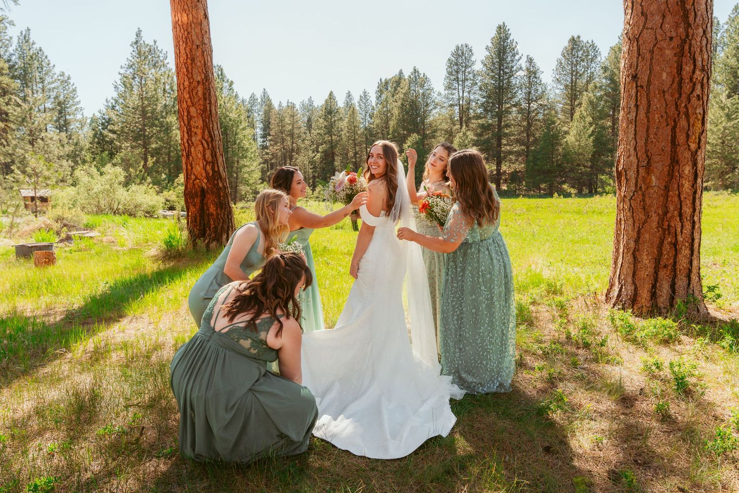 Wedding party poses playfully between tall pine trees in a sunny meadow wearing cream colored attire.
