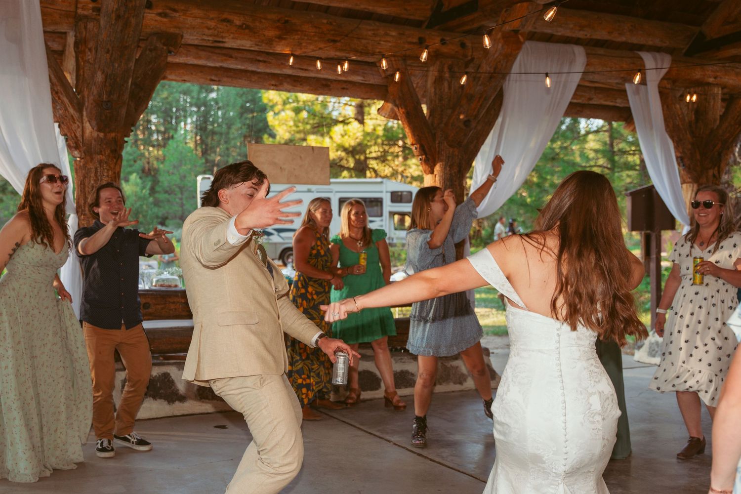 Wedding party dancing under a rustic wooden pavilion during daytime celebration.