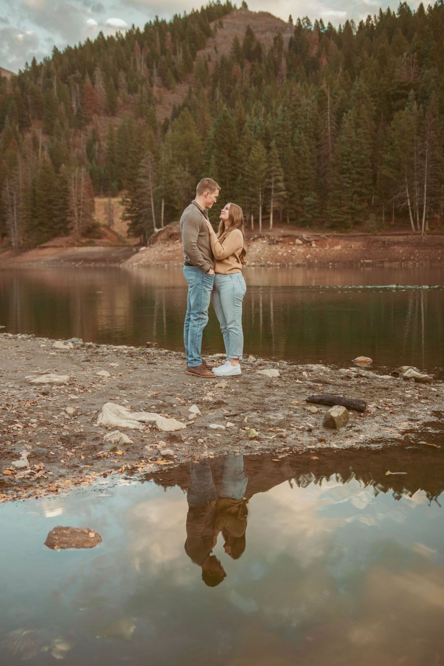Couple standing by a serene mountain lake with their reflection visible in the still water.