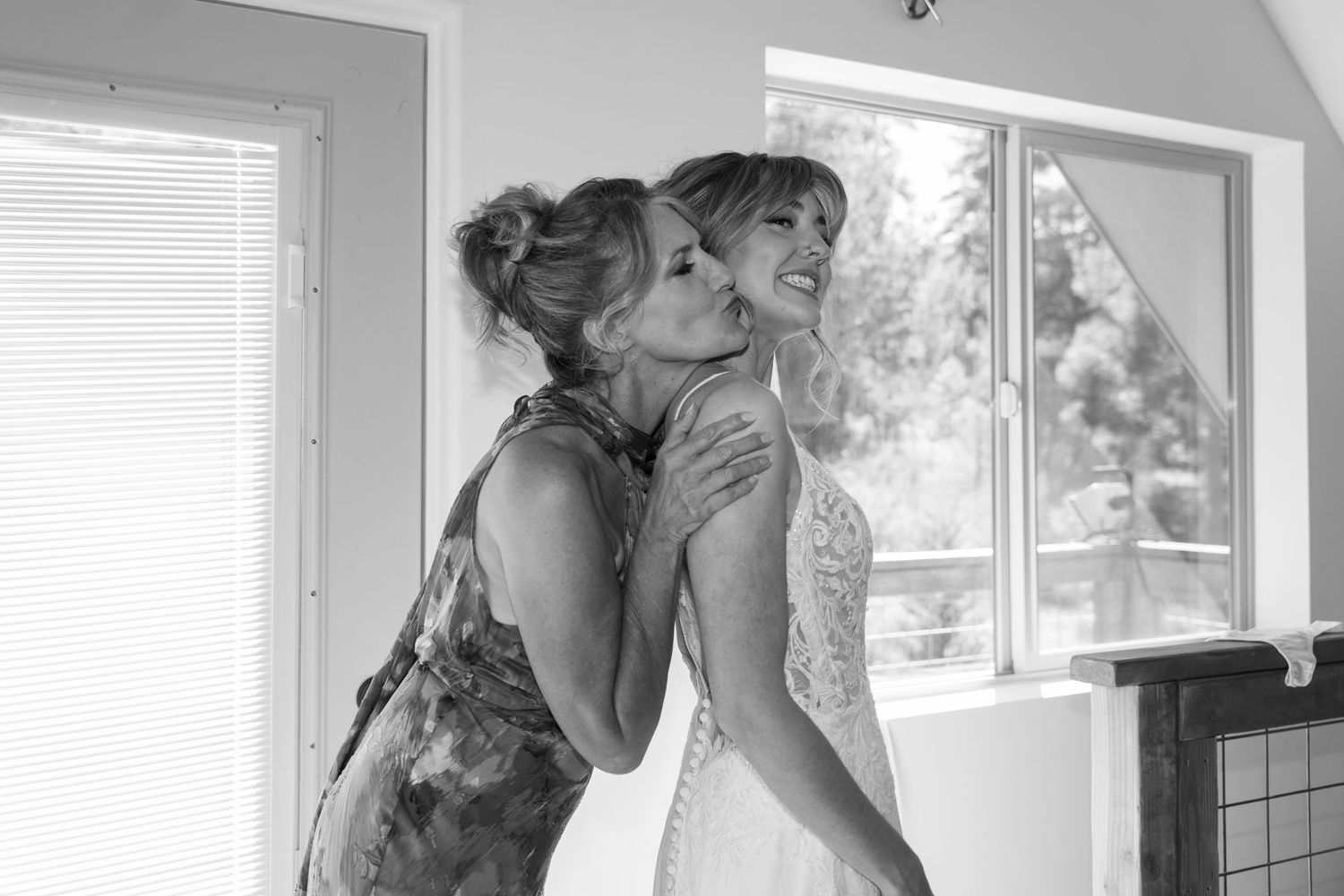 Two people in a bright room, one wearing a floral dress helping another with a white wedding dress.