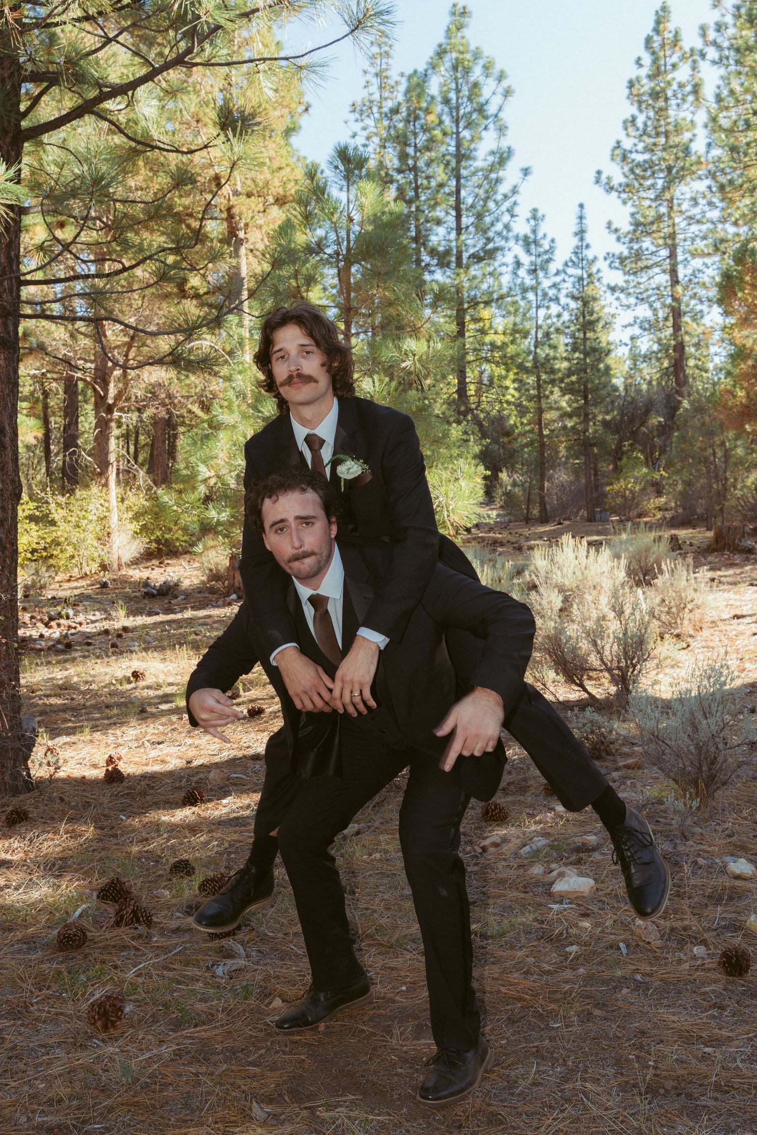 Two people in suits create a playful piggyback pose in a pine forest.