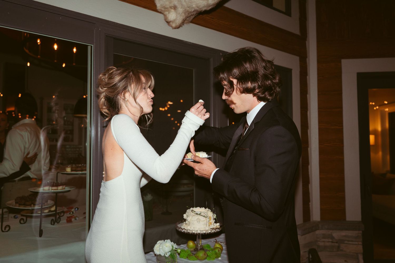 Wedding reception scene with couple sharing cake under indoor lighting.