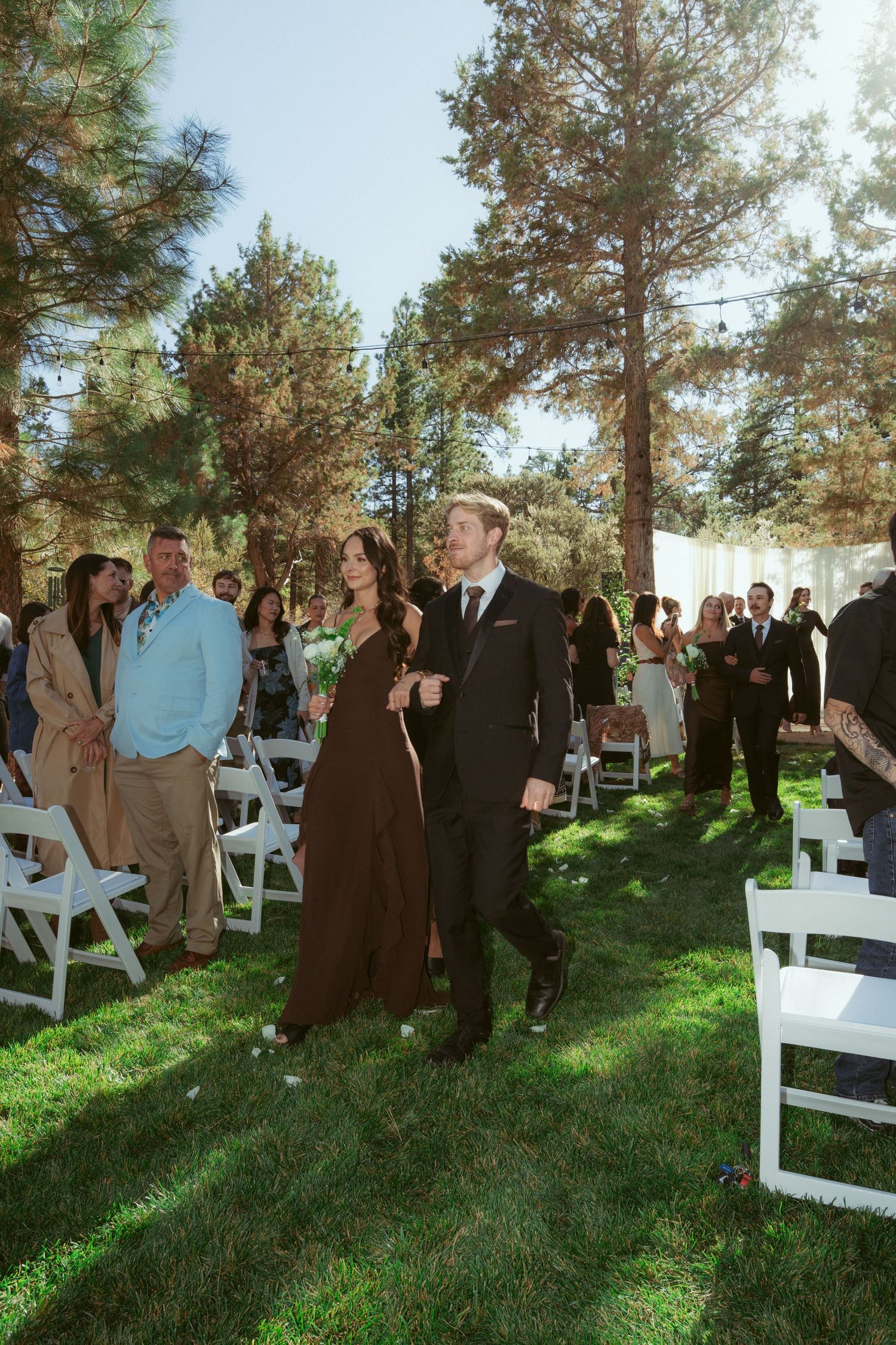 Guests gathering at an outdoor wedding ceremony with white chairs and natural pine backdrop.