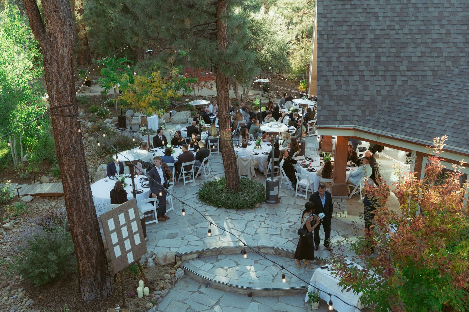 Stone patio wedding venue with white chairs arranged under tall pine trees.