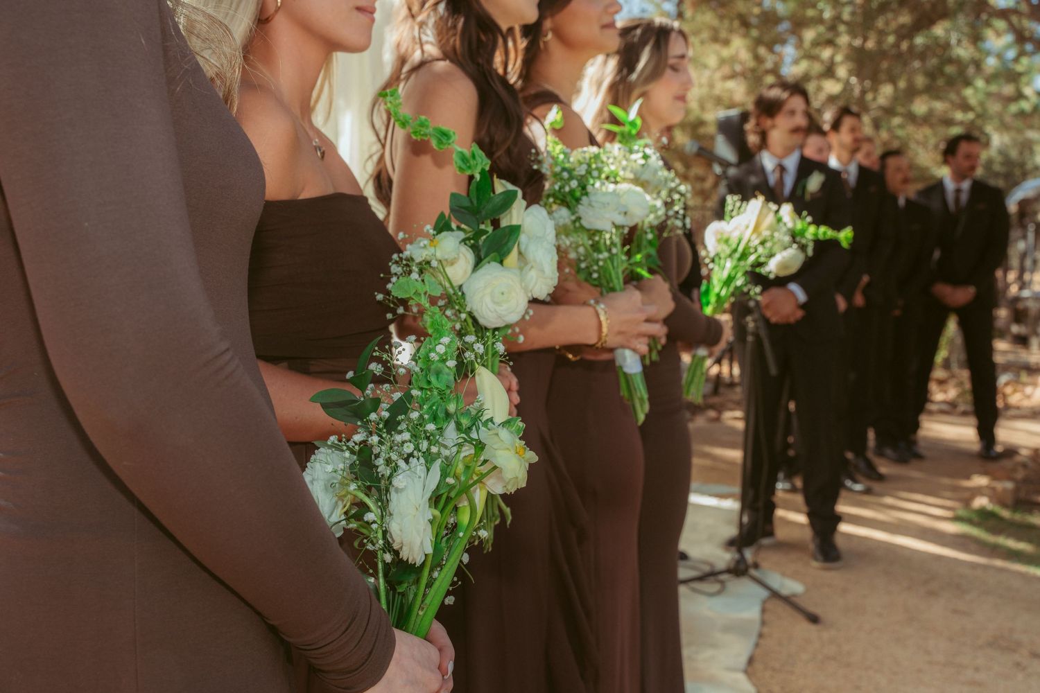 Groomsmen in black suits stand together in a wooded outdoor wedding setting with sunlight filtering through trees.