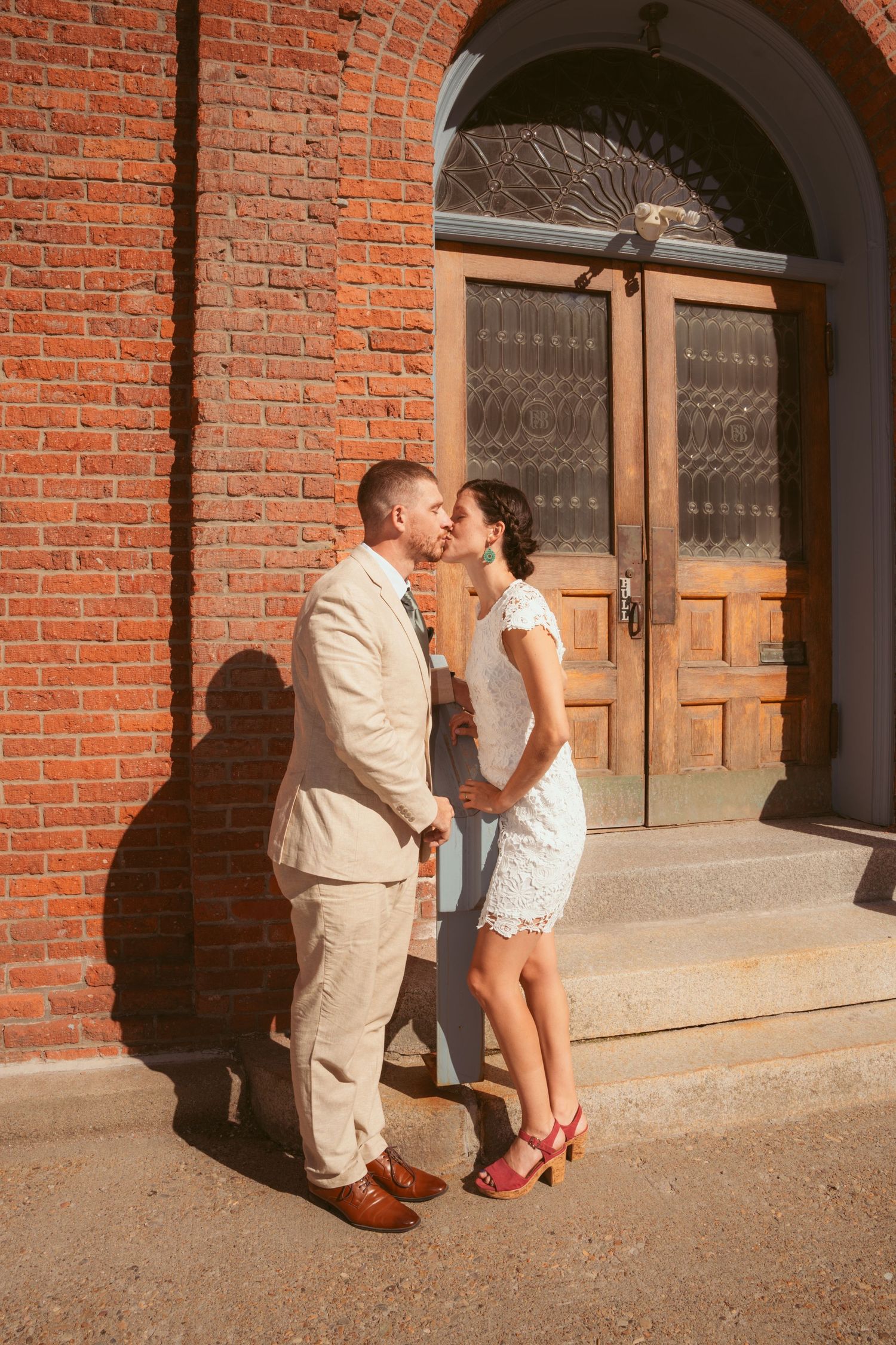 Wedding couple poses playfully against red brick archway in afternoon light.