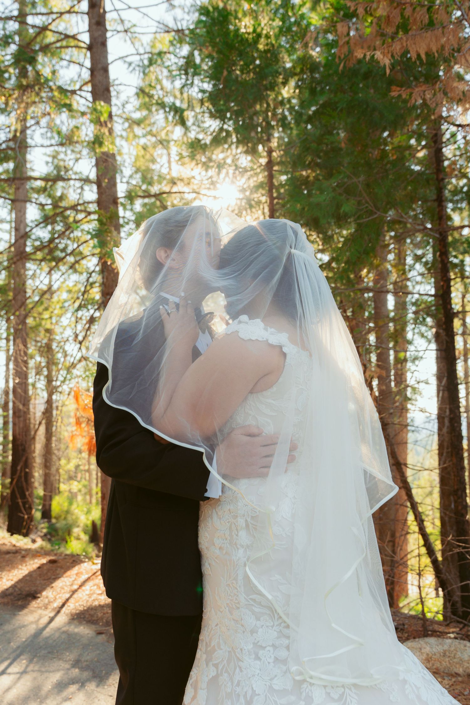 Newlyweds share a romantic moment under sunlit trees with flowing veil.