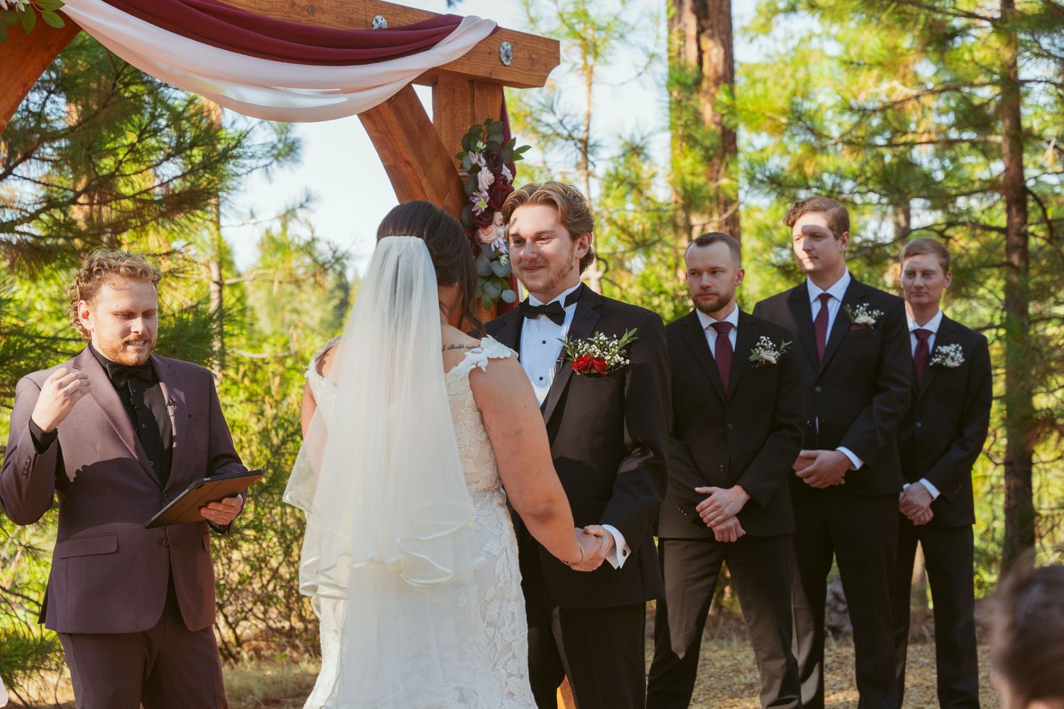 Wedding party standing at altar under pine trees with burgundy draping.