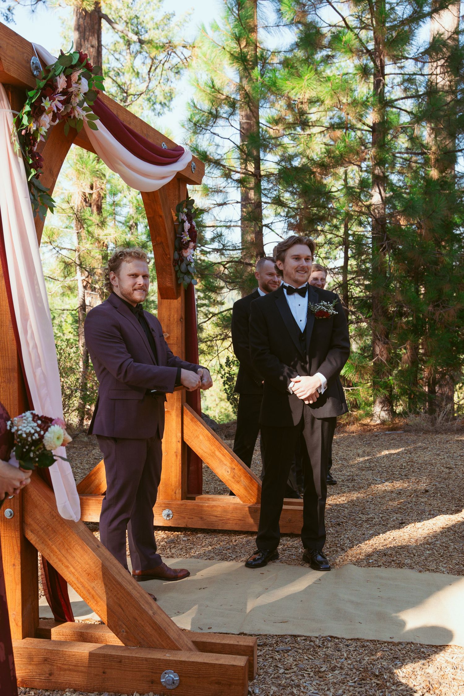 Wedding party members stand at a wooden arch decorated with burgundy draping in a pine forest.