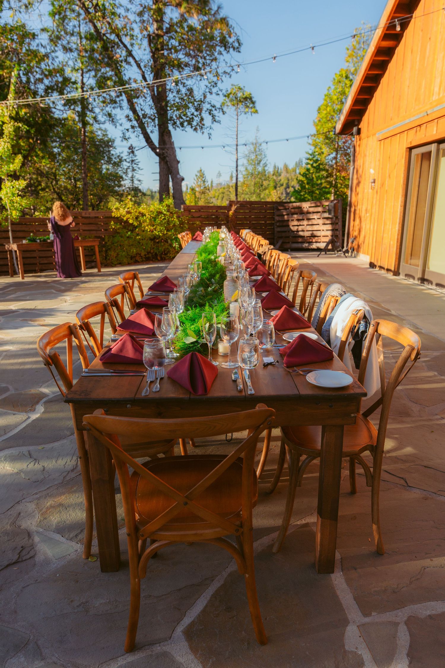 Rustic outdoor wedding reception tables set with burgundy napkins and wooden chairs.