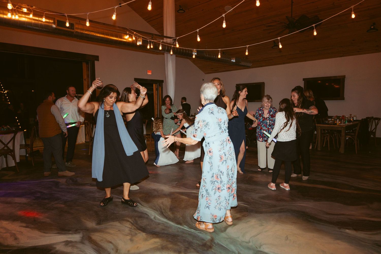 Wedding guests dancing beneath string lights at an indoor reception venue.