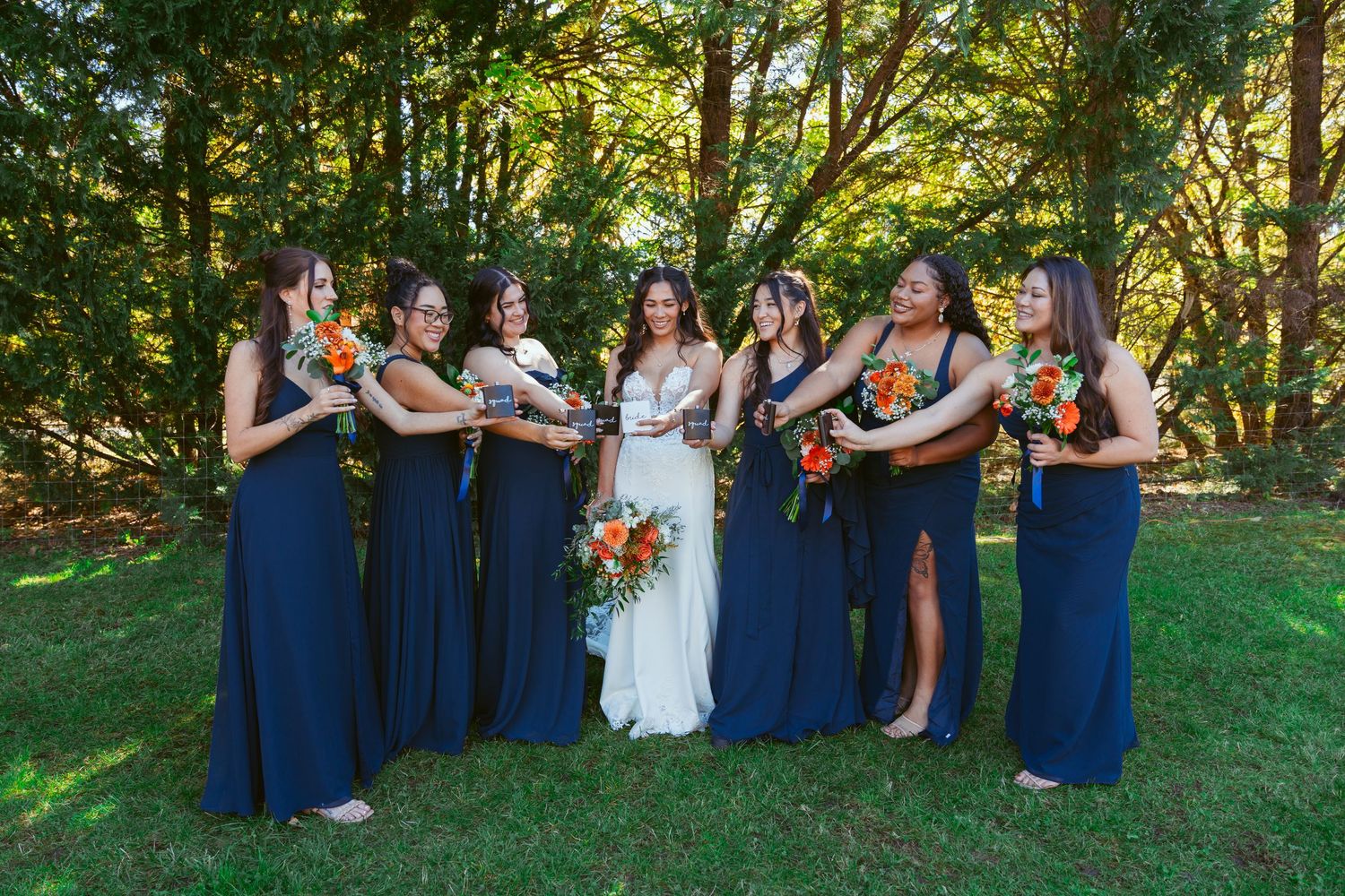 Bridal party in navy dresses laughs together while holding orange bouquets outdoors.