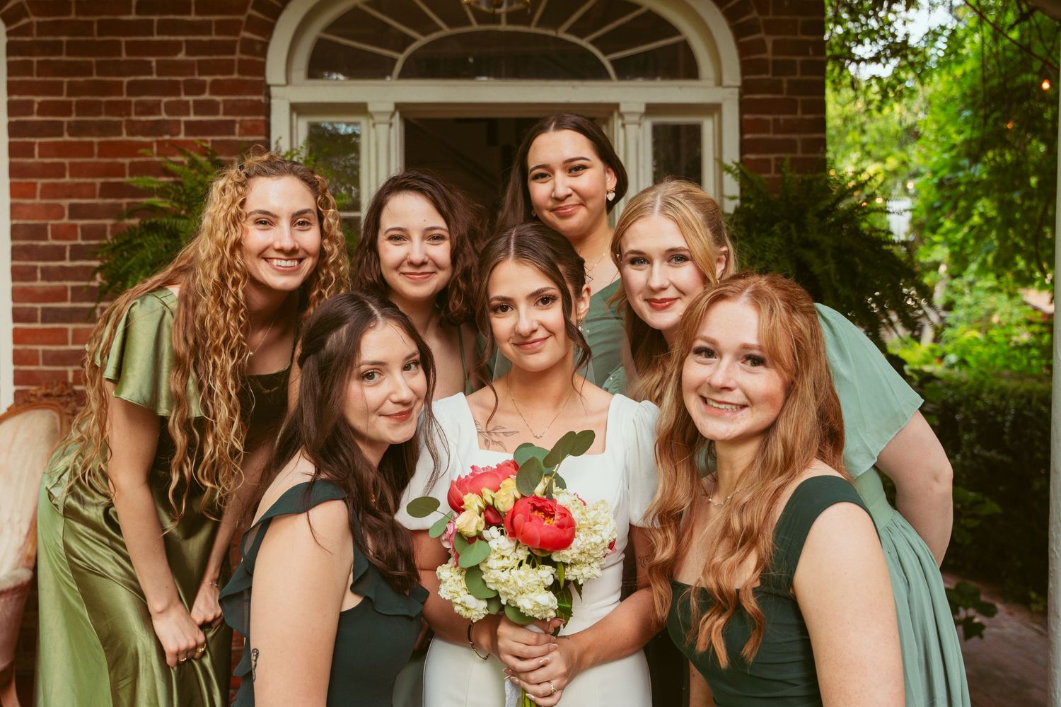 Bridesmaids in sage green dresses gather around bride for group photo at brick entrance.