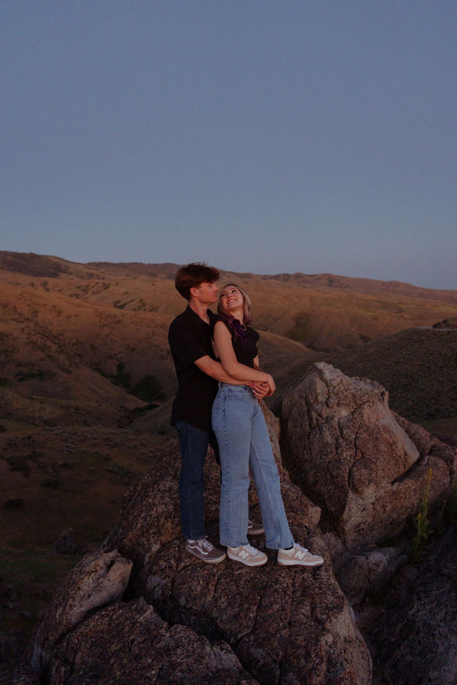 Couple embracing on rocky outcrop with golden hour lighting and mountain views.