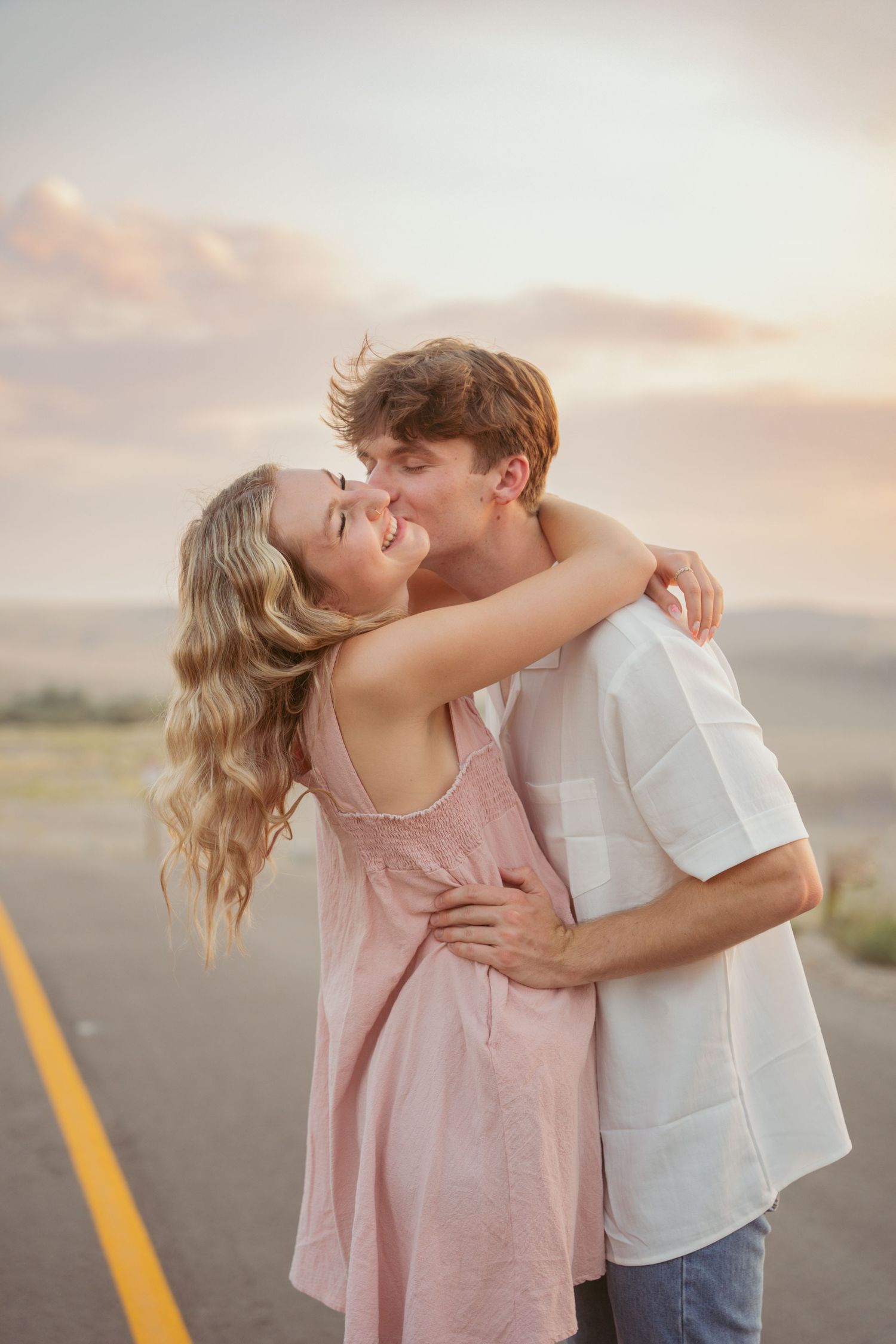 A couple in pink and white outfits share an intimate moment on an empty road at sunset.