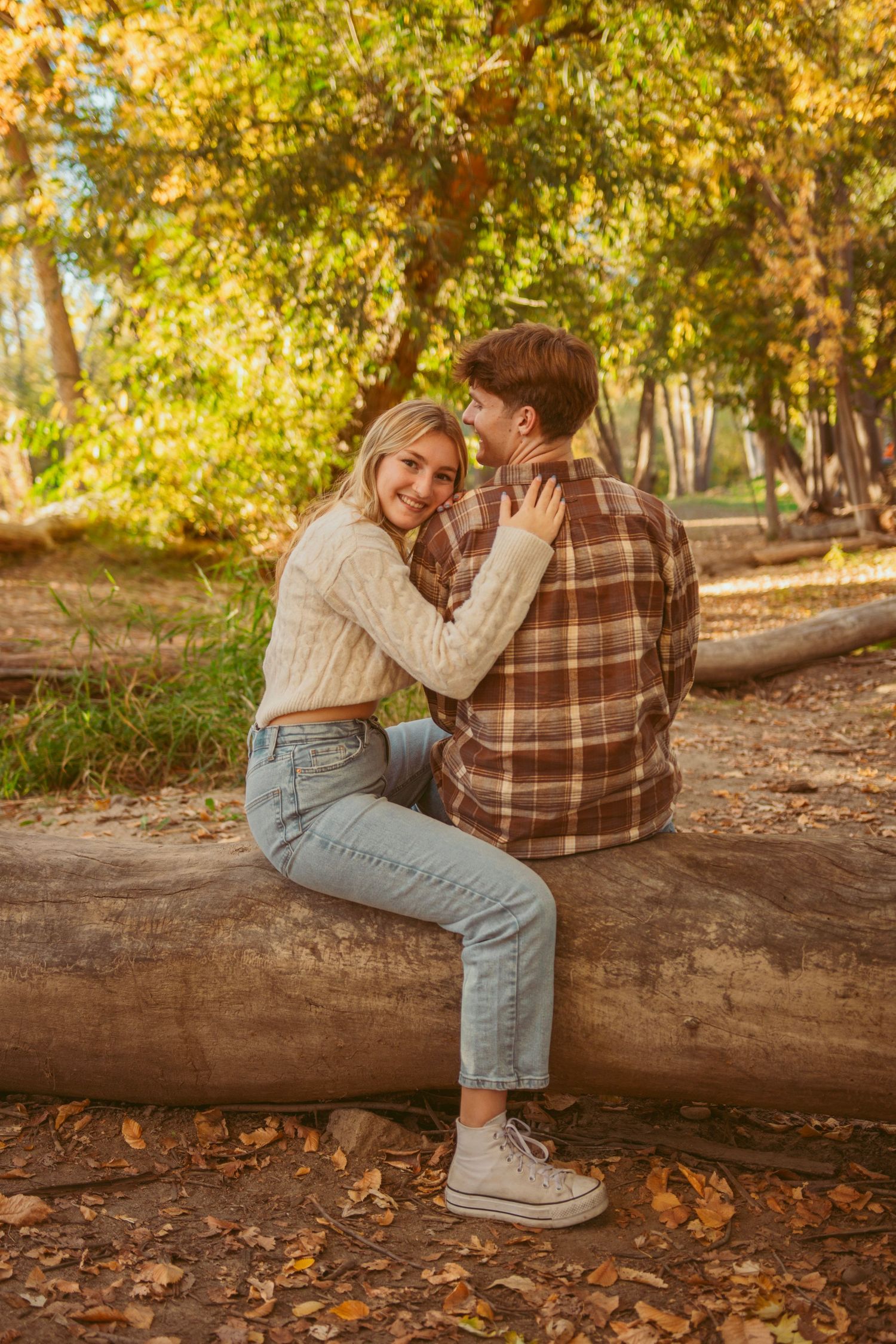 Couple sitting on fallen log surrounded by autumn leaves in park setting.