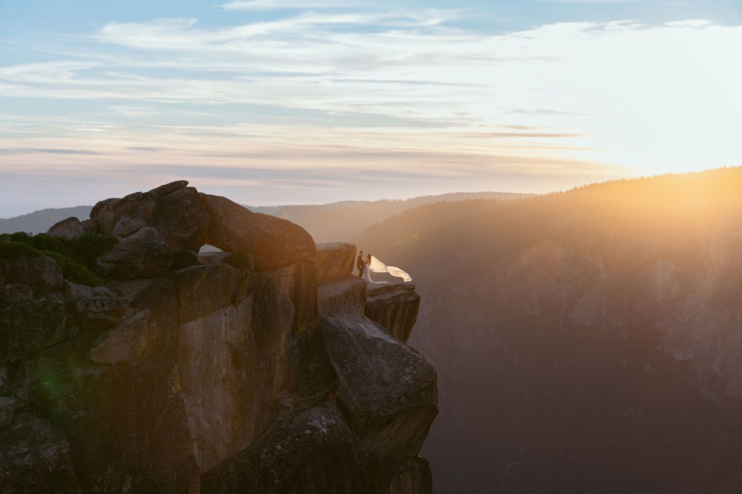 Person sitting on rocky cliff edge at golden sunset with mountain valley views in warm sunlight.