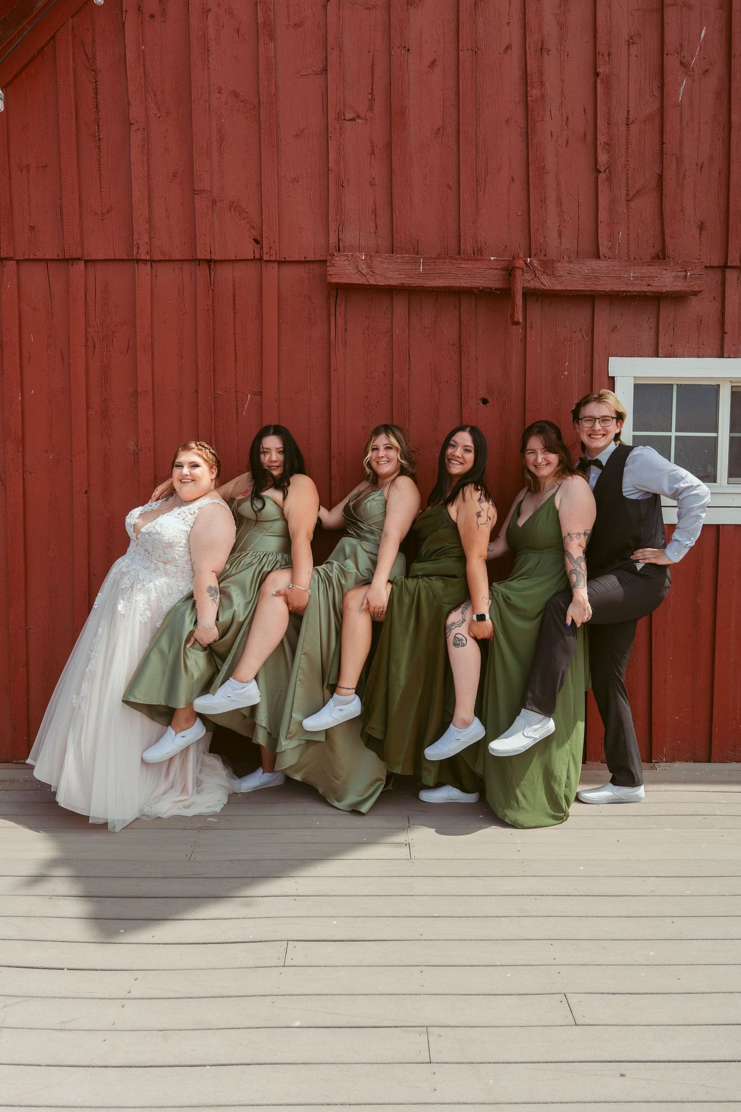 Wedding party in green dresses and white gown pose playfully along deck of red barn.