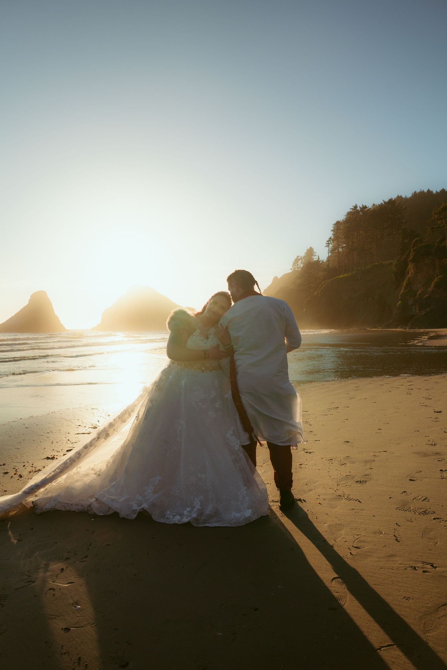 Wedding couple embracing on a beach at sunset with mountains and ocean in the background.