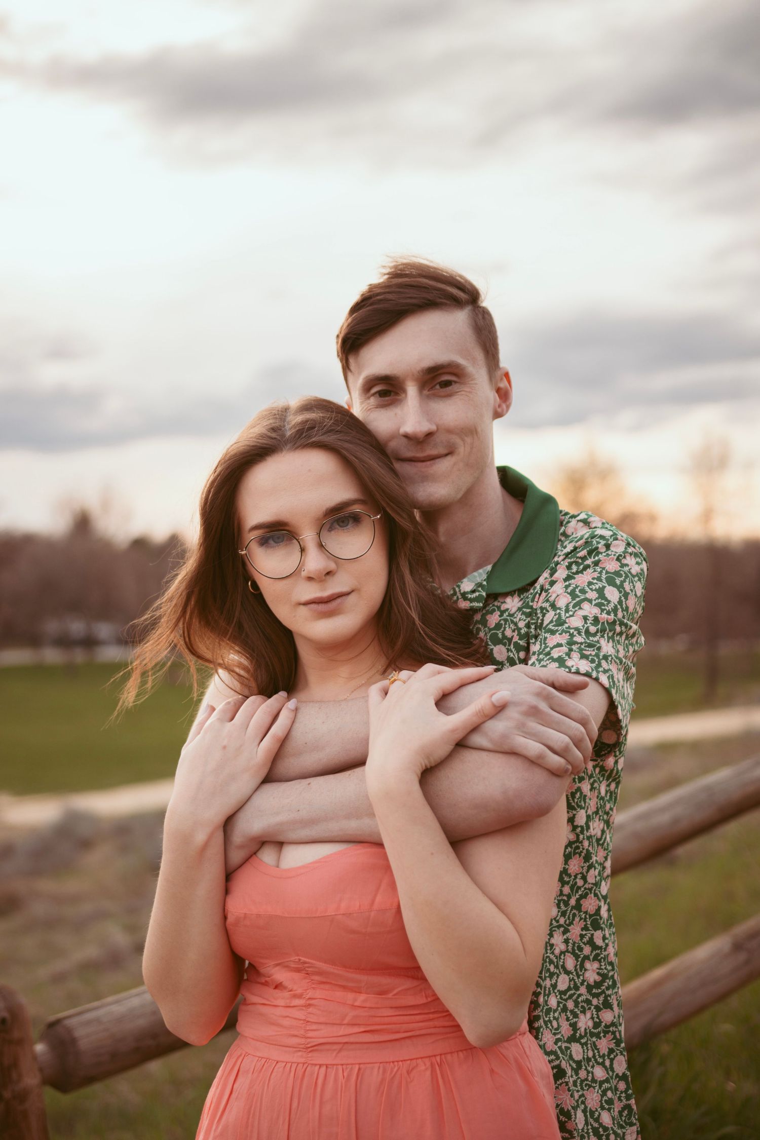 A romantic couple embraces by a wooden fence at sunset with her wearing a coral dress and him in a floral shirt.