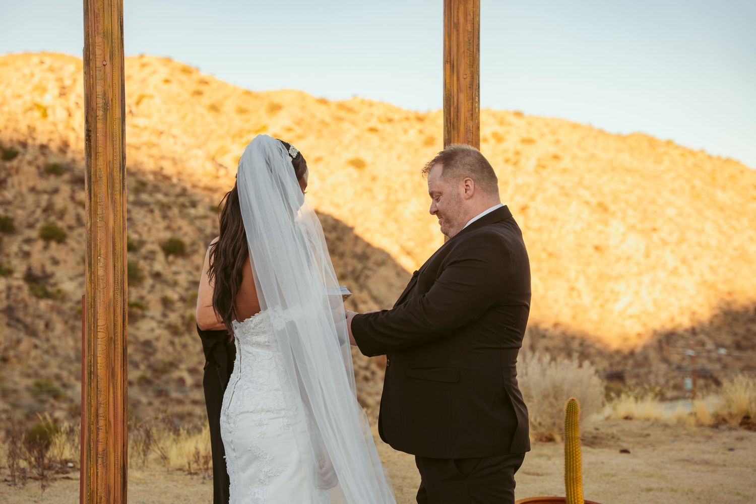 Newlyweds share an intimate moment under a wooden arch with desert mountains in the background.