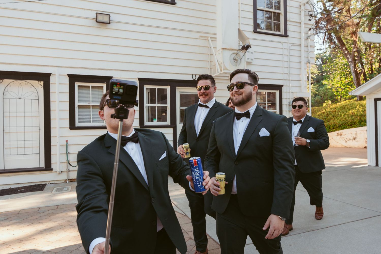 Groomsmen in black tuxedos and sunglasses pose outside white building.