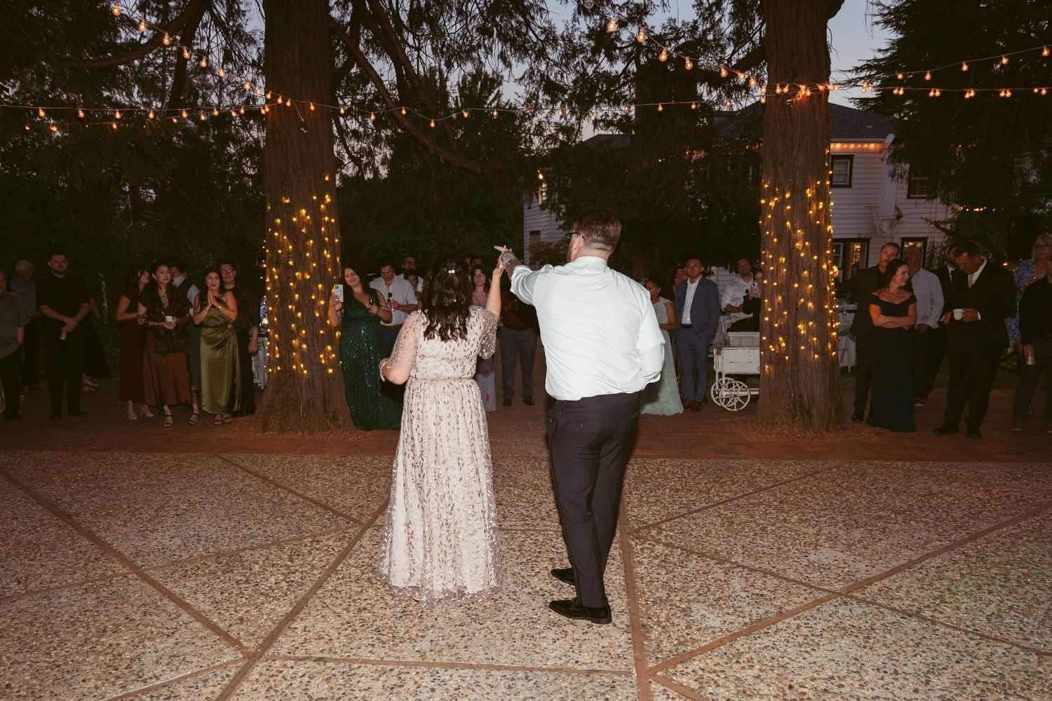Couples dancing at dusk under illuminated trees at an outdoor wedding celebration.