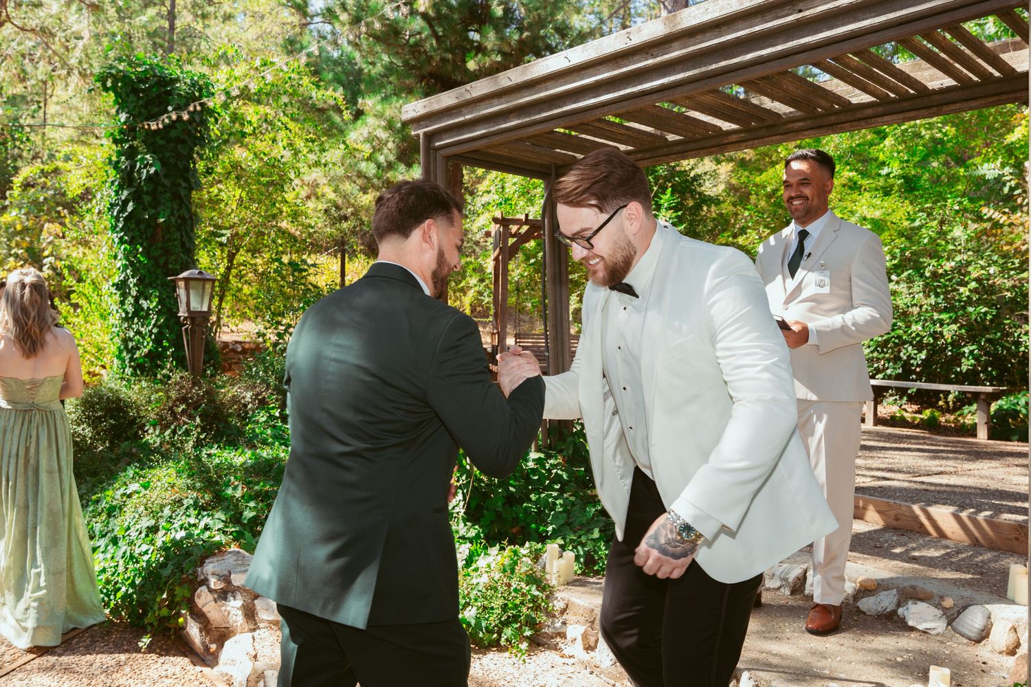 Two people in formal attire shake hands during an outdoor wedding ceremony under a wooden pergola.