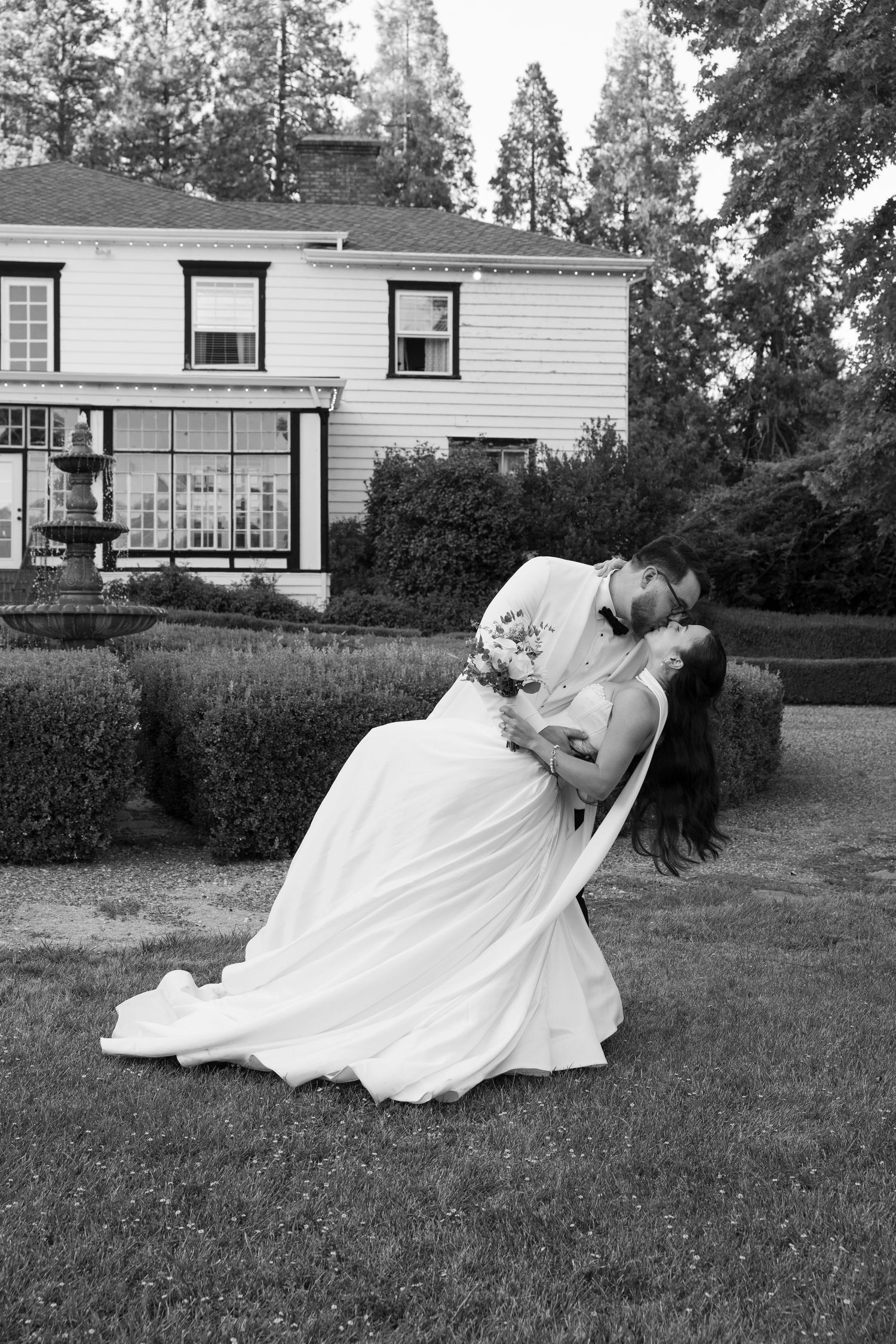 A couple in wedding attire dips gracefully on a grassy lawn in front of a white house in black and white.