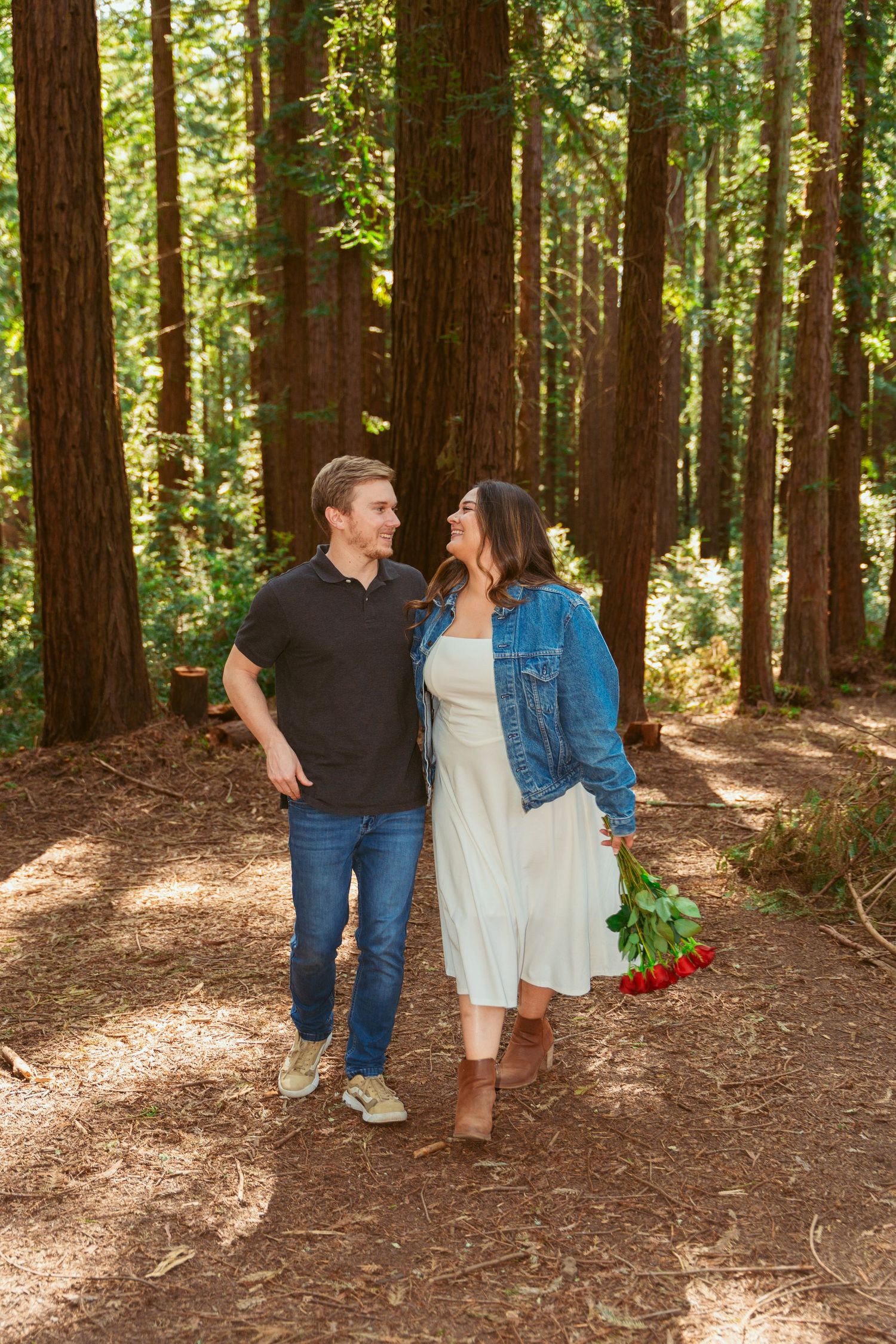Two people stroll together through a peaceful redwood grove holding hands and carrying flowers.