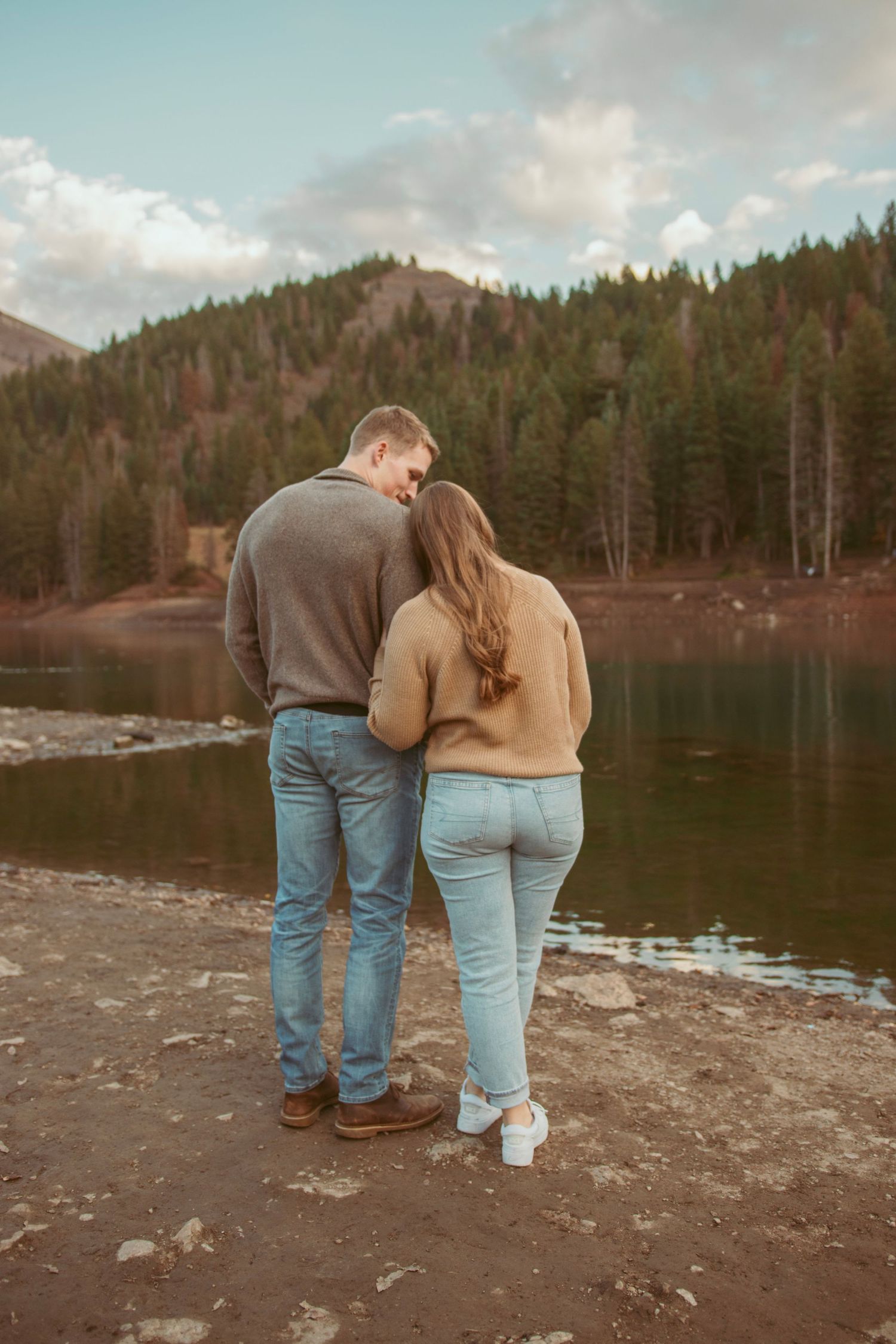 Couple standing close together on the shore of a mountain lake during sunset.