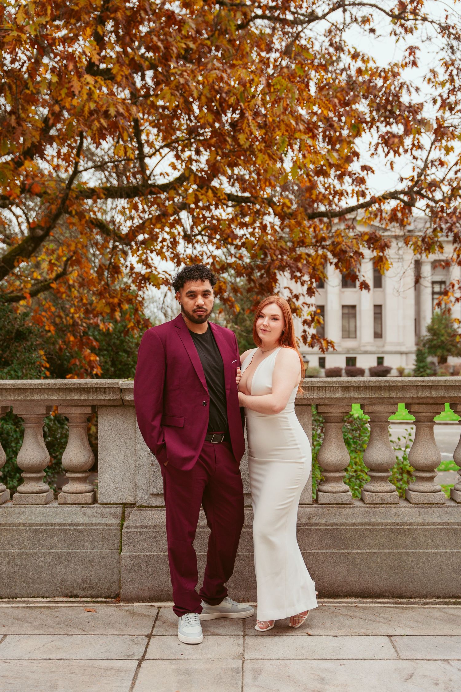 A formal portrait of a couple against autumn foliage and classical architecture.