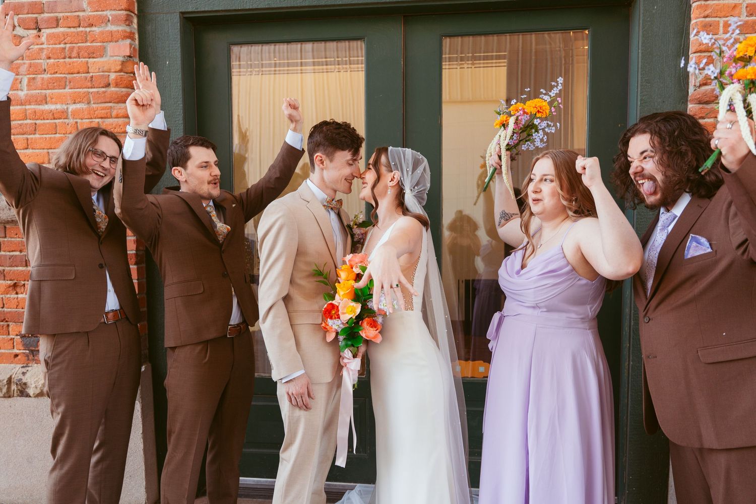 Wedding party poses along brick wall in purple bridesmaid dresses and brown groomsmen suits.
