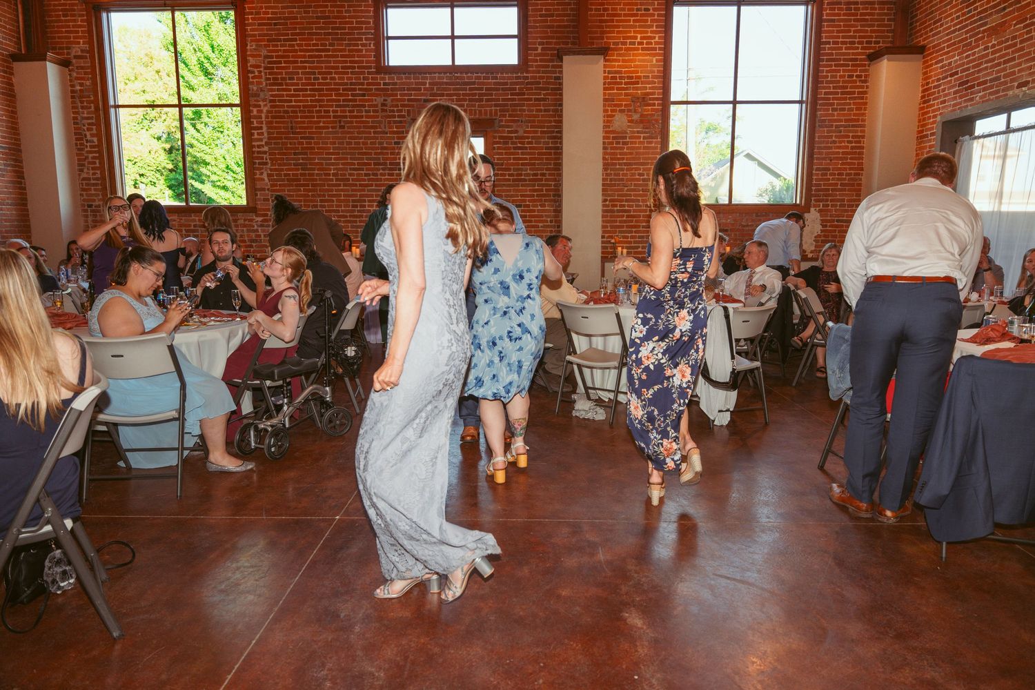Wedding guests dance and celebrate on the dance floor of a rustic brick venue with large windows.