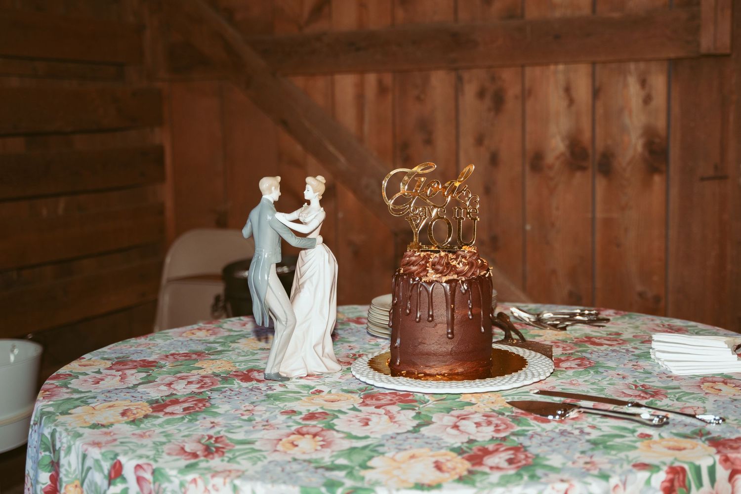 Wedding cake table featuring a chocolate tiered cake and porcelain topper on floral patterned tablecloth.