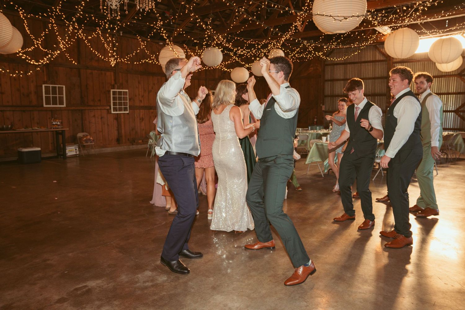 Wedding party dances together under warm string lights and lanterns in a rustic barn setting.