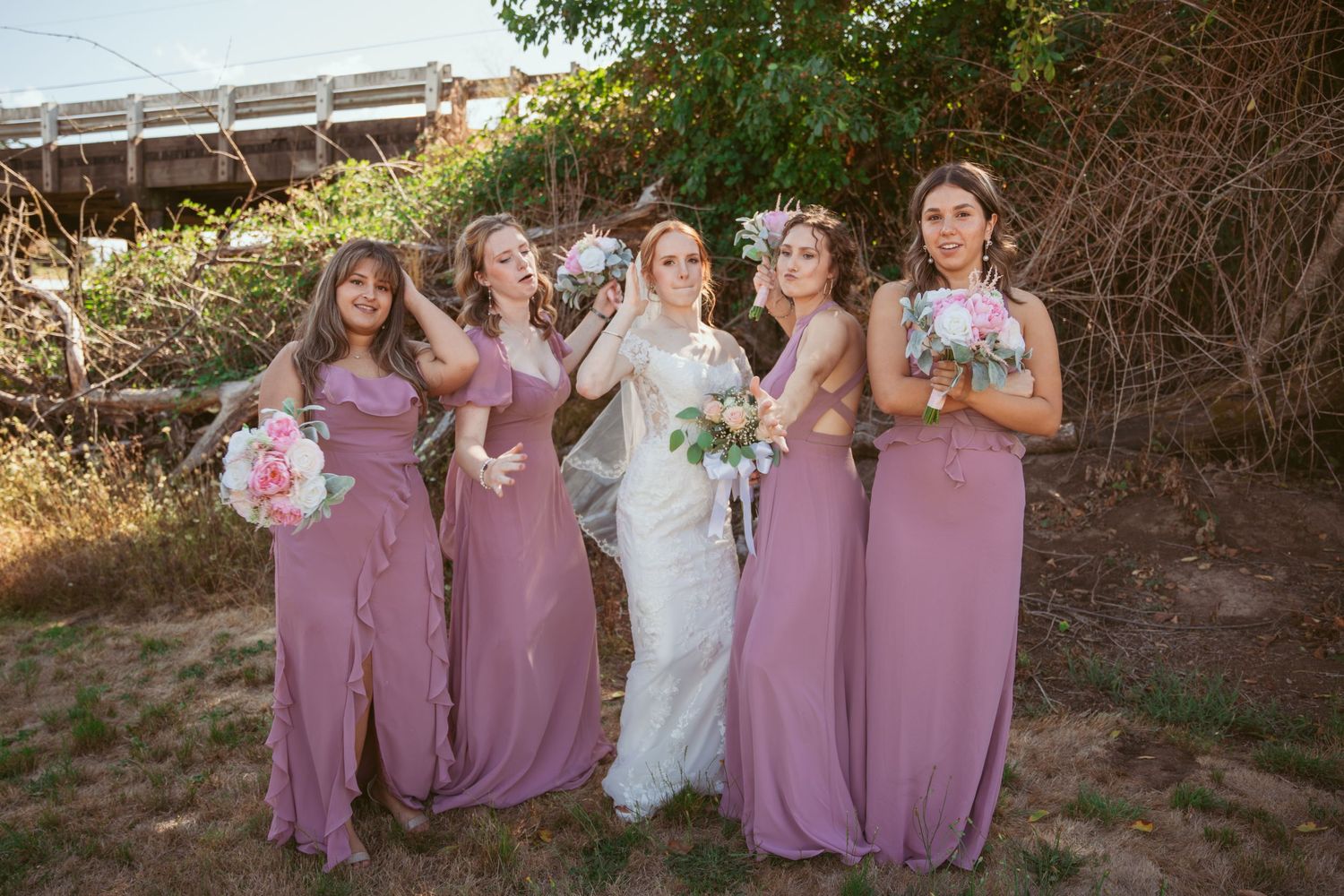 Bridesmaids in dusty rose dresses pose together with bride in white gown against natural outdoor backdrop.